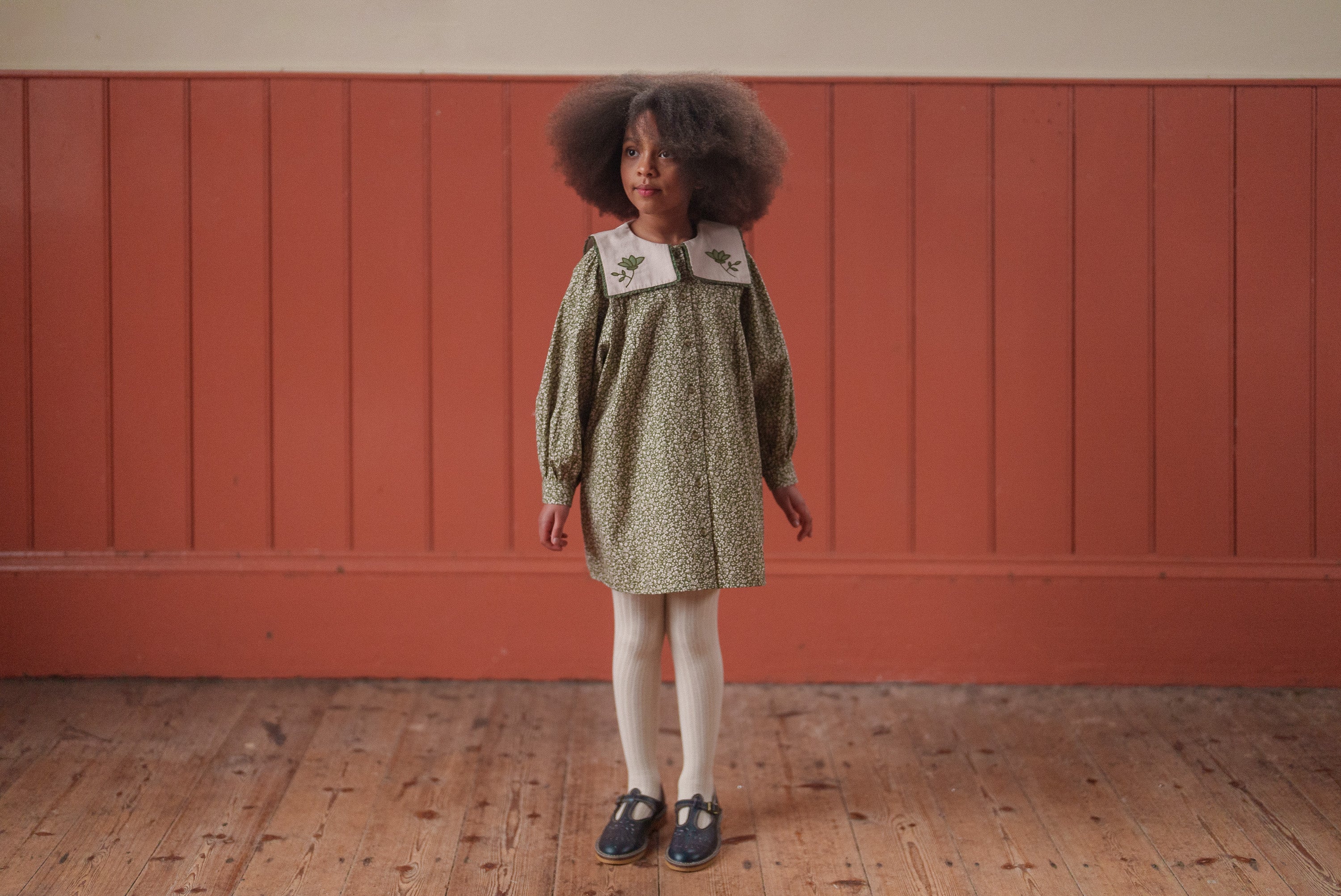 Young girl wearing a green floral organic cotton dress with a vintage style collar, standing on a wooden floor against a muted terracotta wall in sustainable children's clothing.