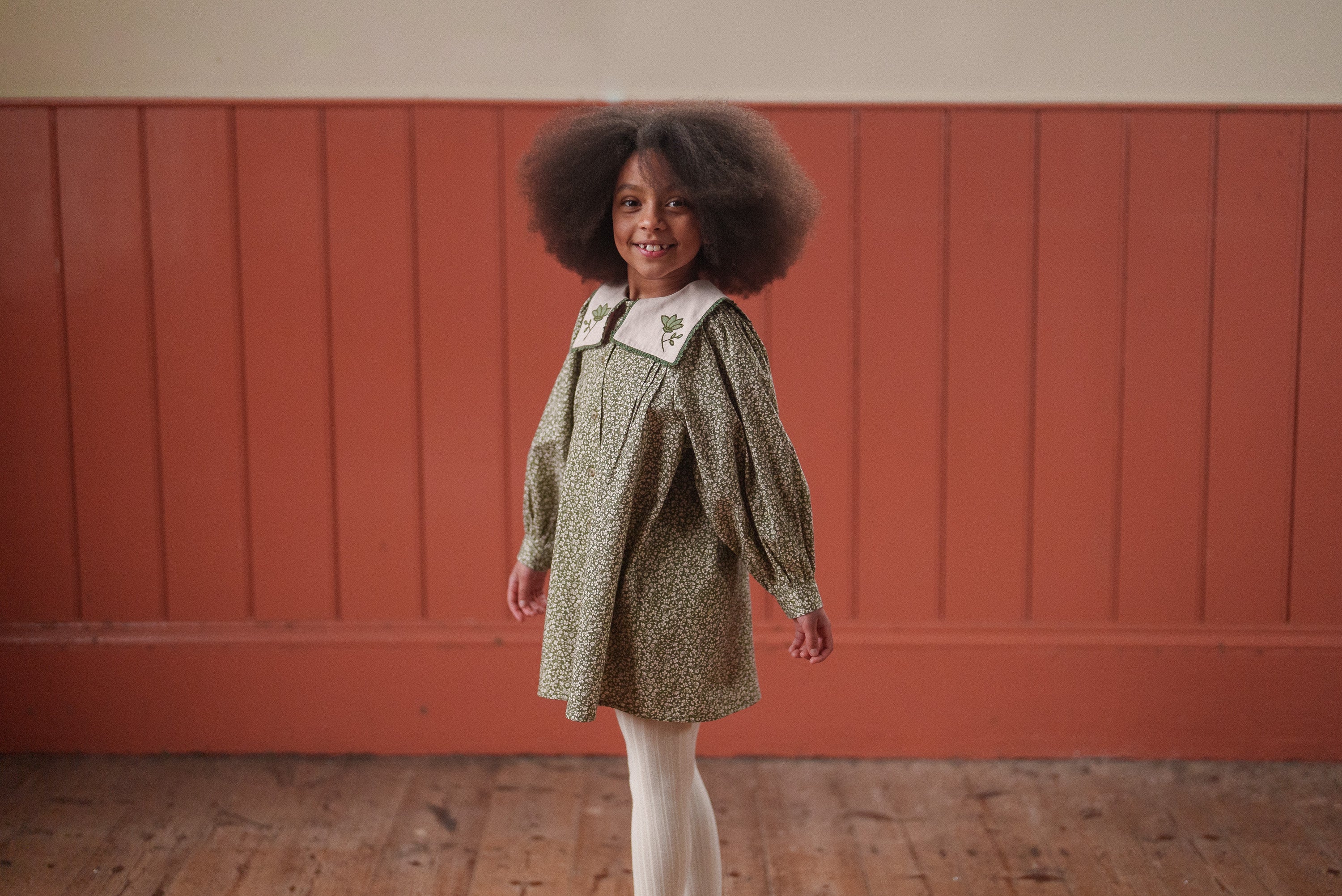 A young girl wearing a vintage style green floral organic cotton dress with a white embroidered collar, posing indoors against a warm-toned wooden wall.