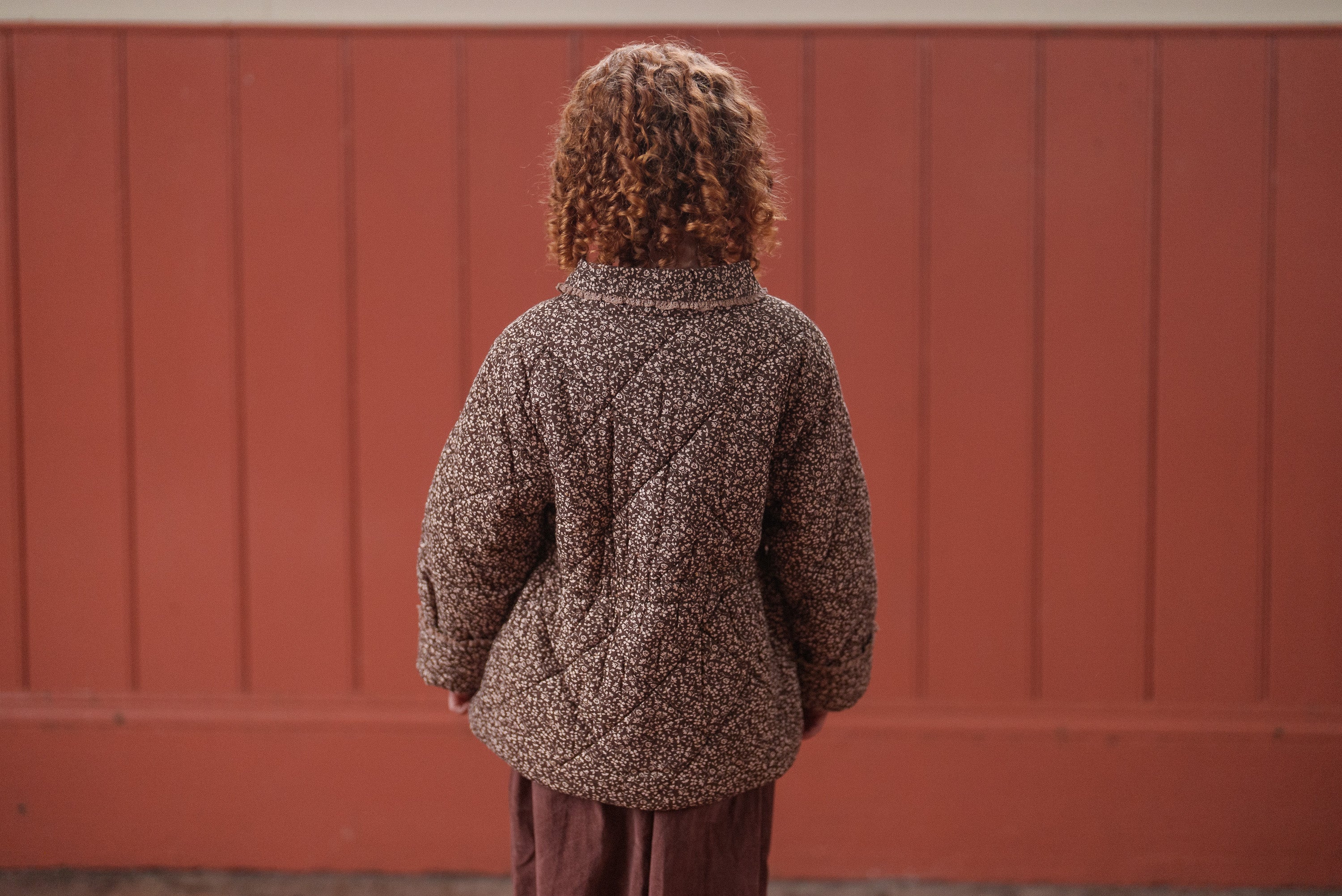 Child wearing a brown and beige floral organic cotton jacket with a quilted design, standing against a muted red wooden wall, showcasing sustainable vintage style kids clothing.