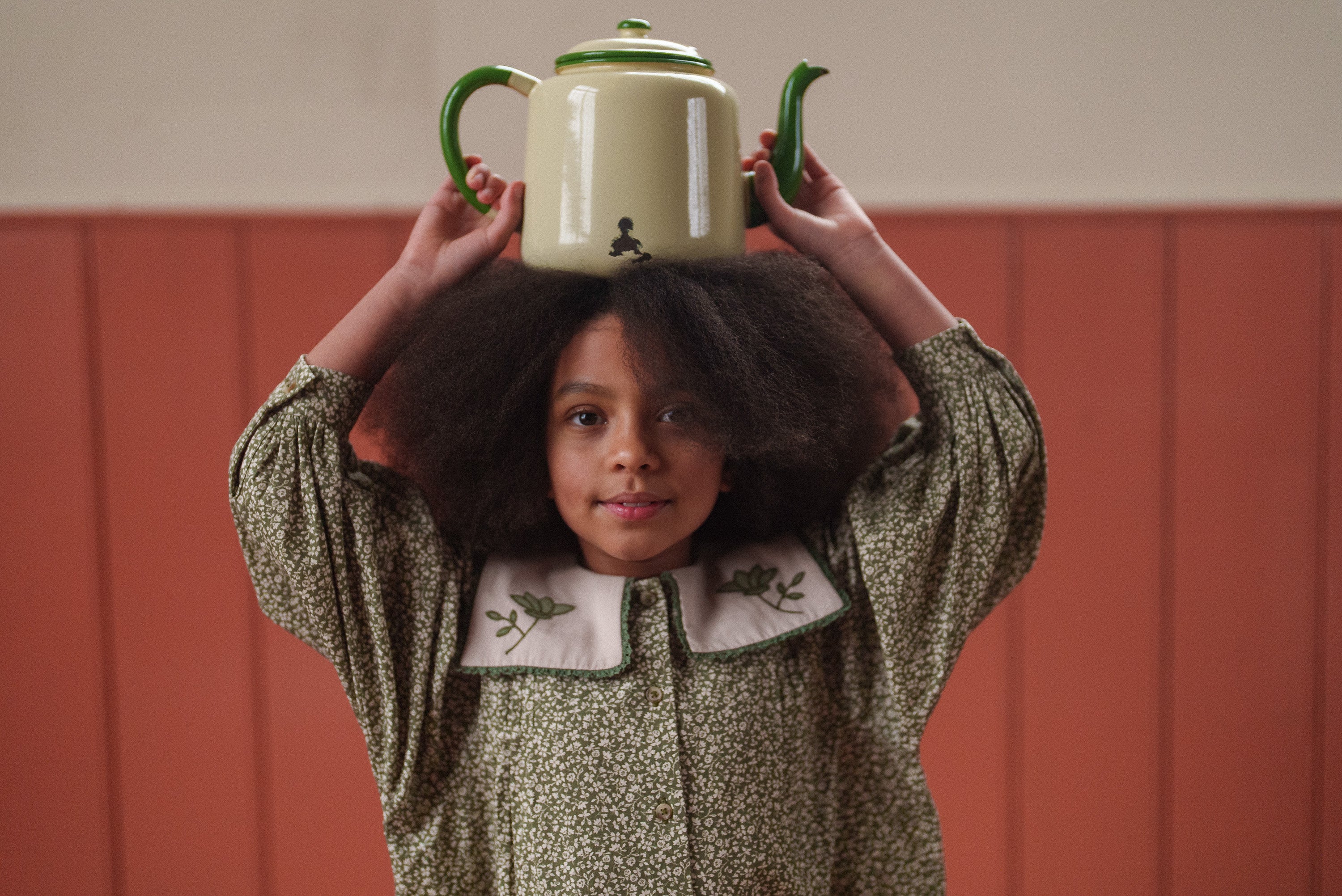 Young girl wearing an organic cotton vintage-style green floral dress with embroidered collar, balancing a cream and green teapot on her head, showcasing sustainable kids clothing.