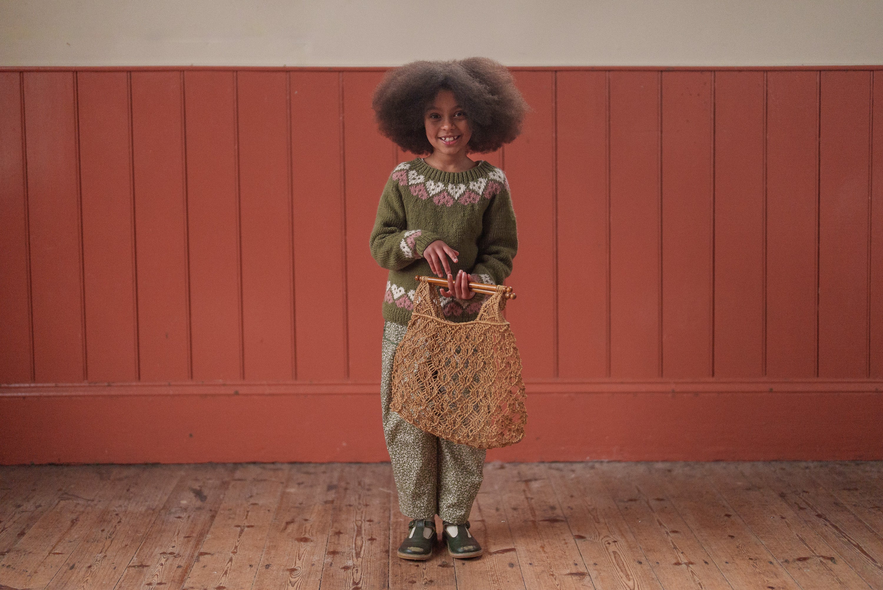 Young girl wearing a hand-knitted multicolor merino wool jumper with a vintage style, holding a natural woven bag in a warm, rustic room setting.