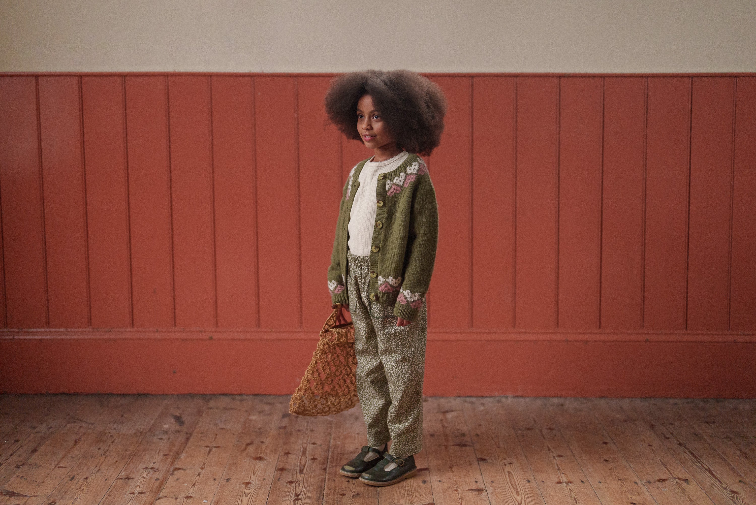 Child wearing a multicolor blue hand-knitted merino cardigan with hearts over vintage style trousers, holding a woven bag in a natural light setting