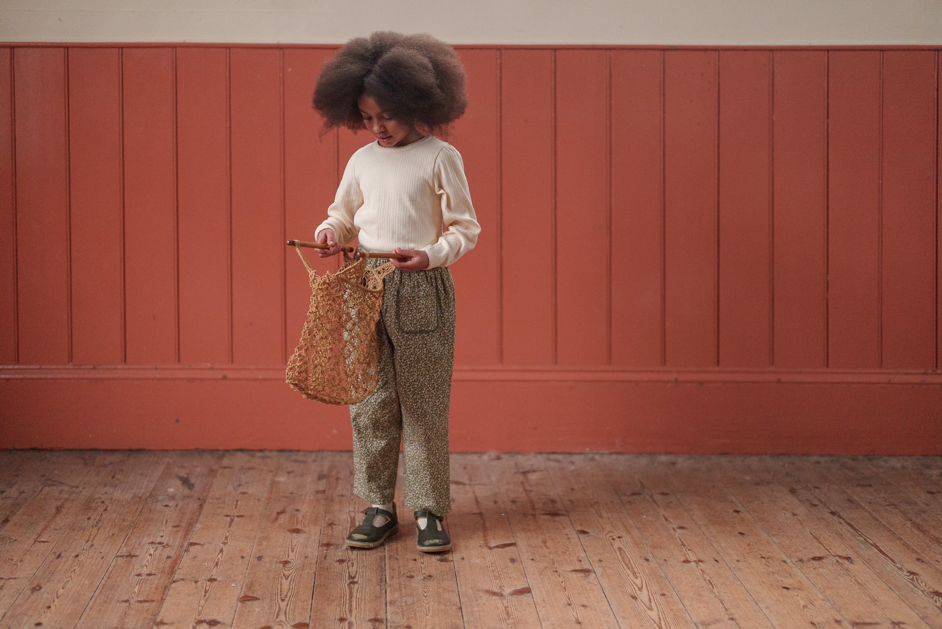 Young child wearing vintage style organic cotton green floral trousers and cream sweater, holding woven bag in natural wooden floor room with warm orange wall.