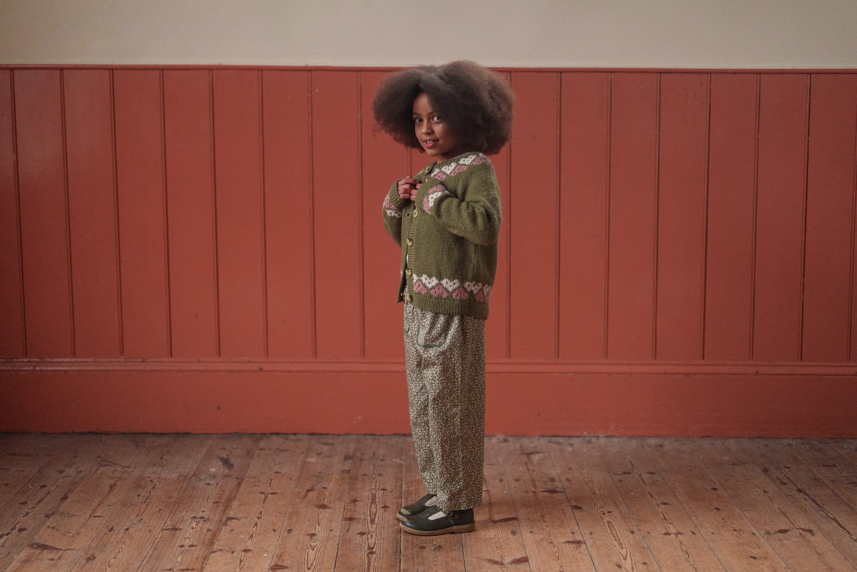 Young child wearing a vintage-style hand-knitted wool cardigan in blue and multicolor over patterned pants, standing on wooden floor against a warm-toned paneled wall in natural, ethical kids clothing.