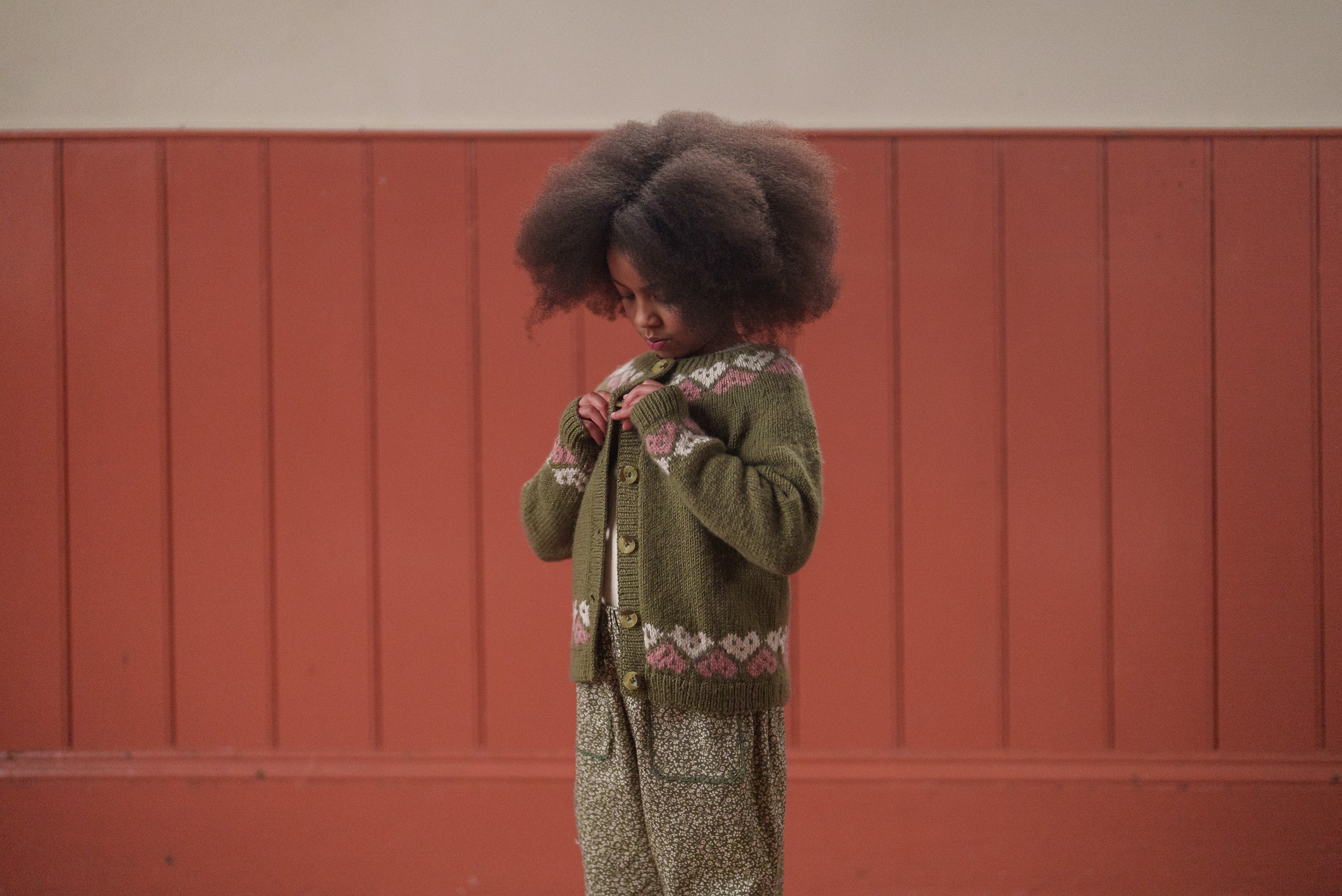 Child wearing a vintage style, hand-knitted multicolor merino cardigan with heart patterns, styled with loose patterned pants in a natural, ethical children's clothing setting.