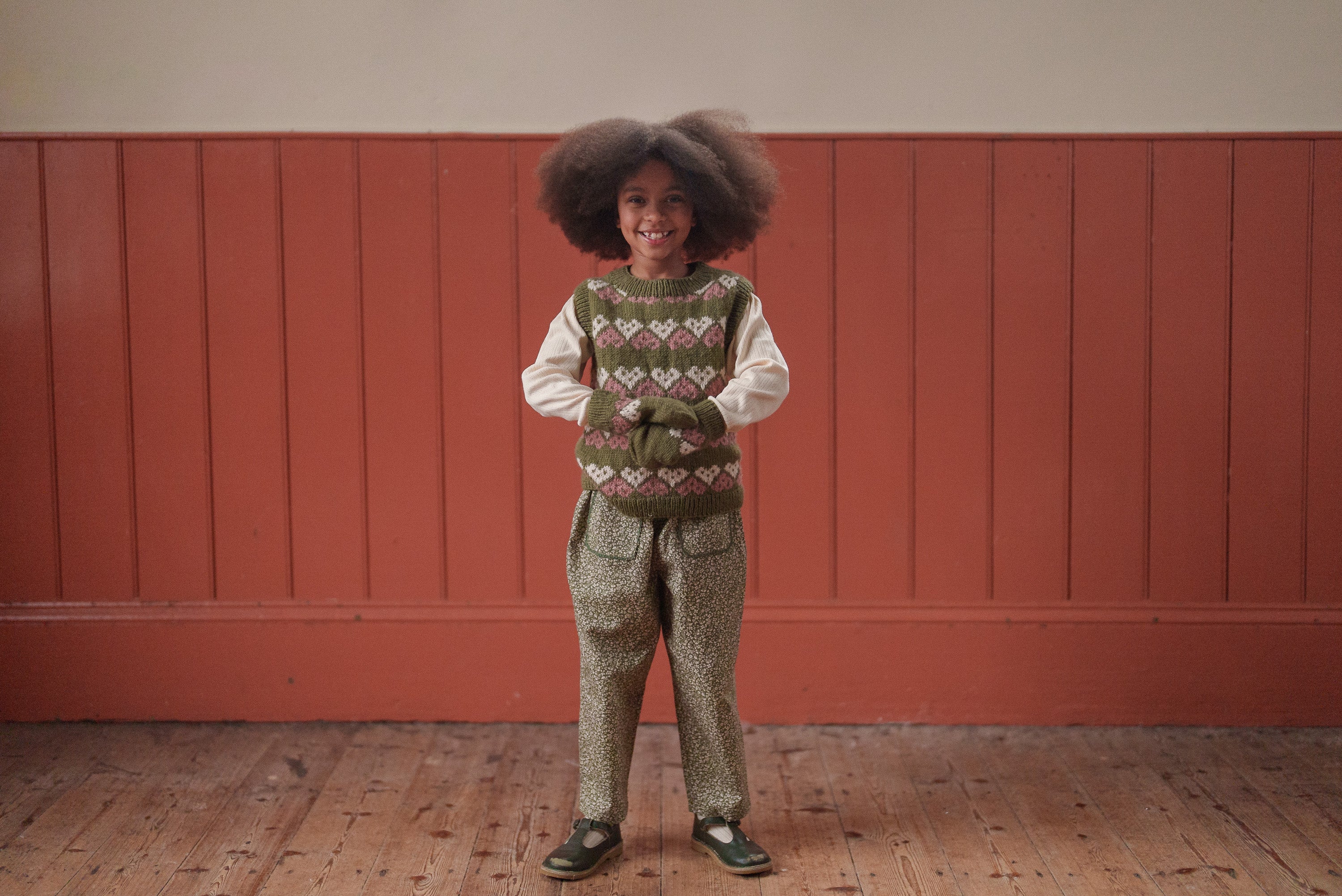 Smiling child wearing vintage style multicolor hand-knitted heart mittens and patterned trousers, showcasing sustainable, ethical children’s clothing in natural materials.