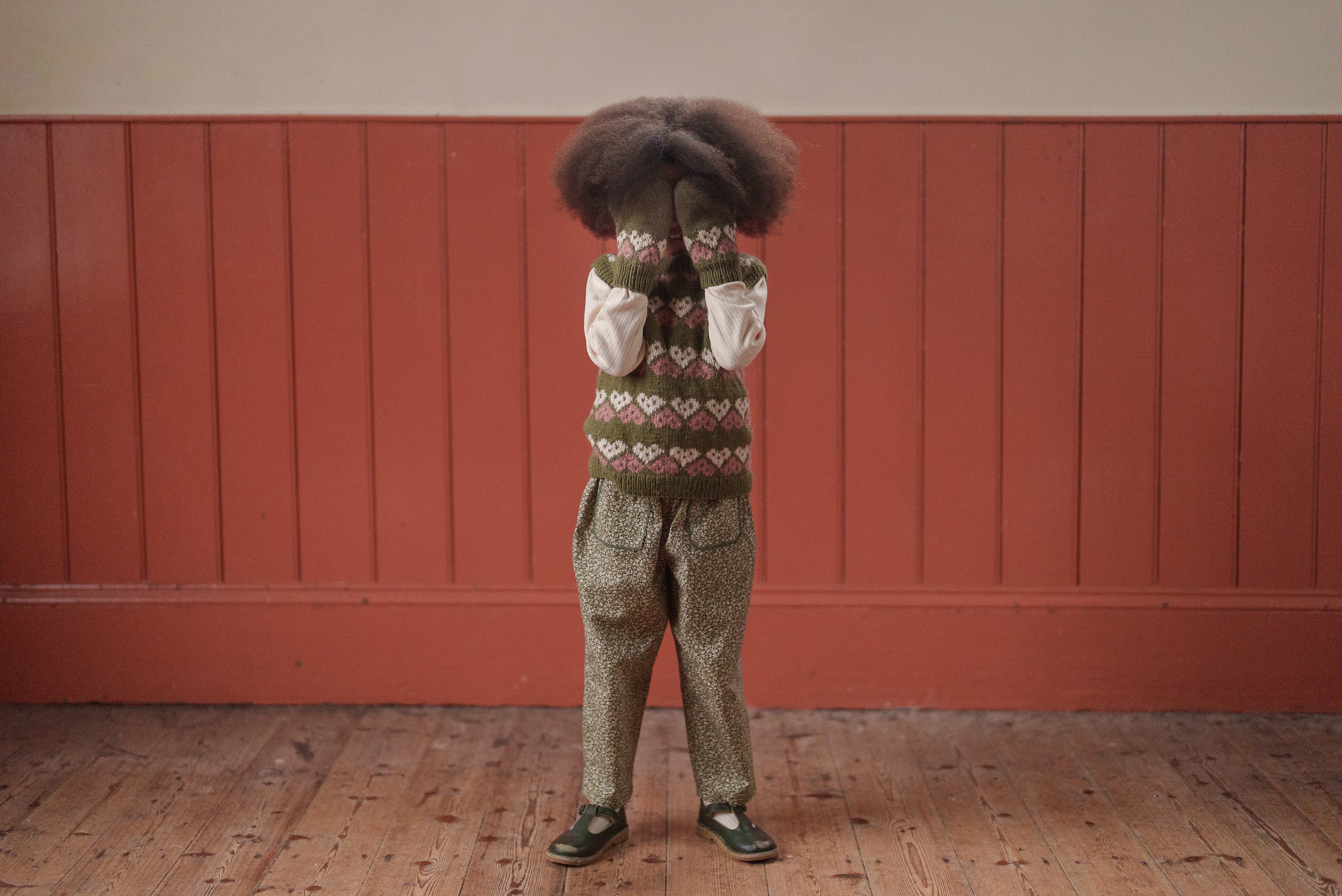 Child wearing vintage style green floral organic cotton trousers paired with a patterned vest and white long-sleeve top in a natural, slow fashion setting.