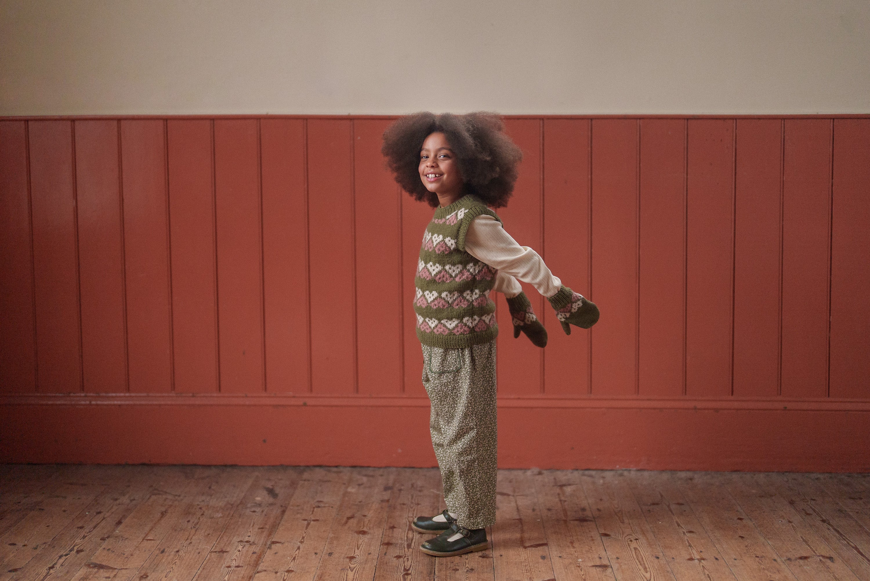 A happy child wearing green floral organic cotton trousers and a patterned knitted vest, styled with vintage-inspired sustainable kids clothing in a warm indoor setting.