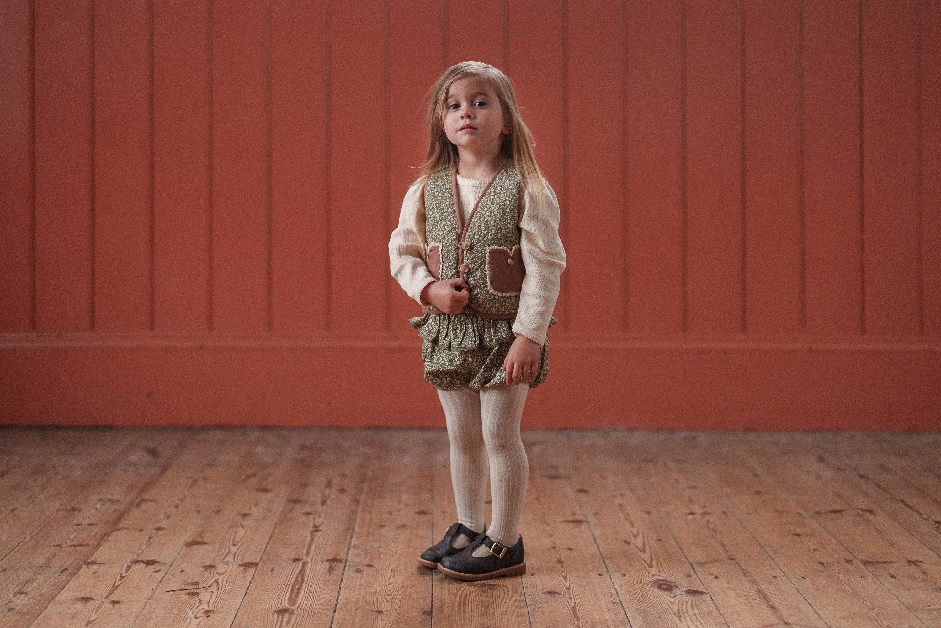 Young girl wearing a vintage-style organic cotton romper with a green floral pattern, paired with cream-colored tights and black shoes, standing on a wooden floor.