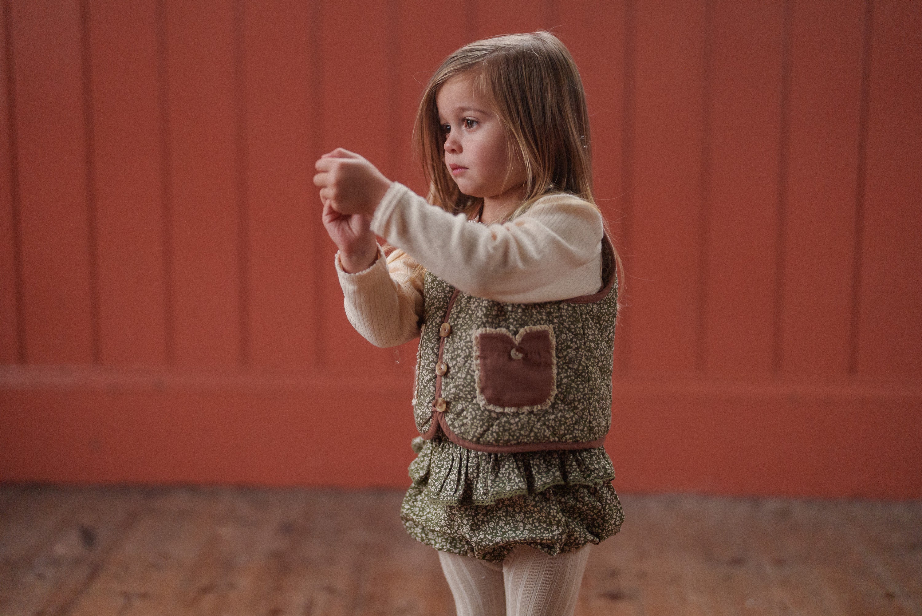 Young girl wearing a vintage style frilly romper in green floral organic cotton with a cream long-sleeve top, showcasing slow, natural, and ethical children's clothing.