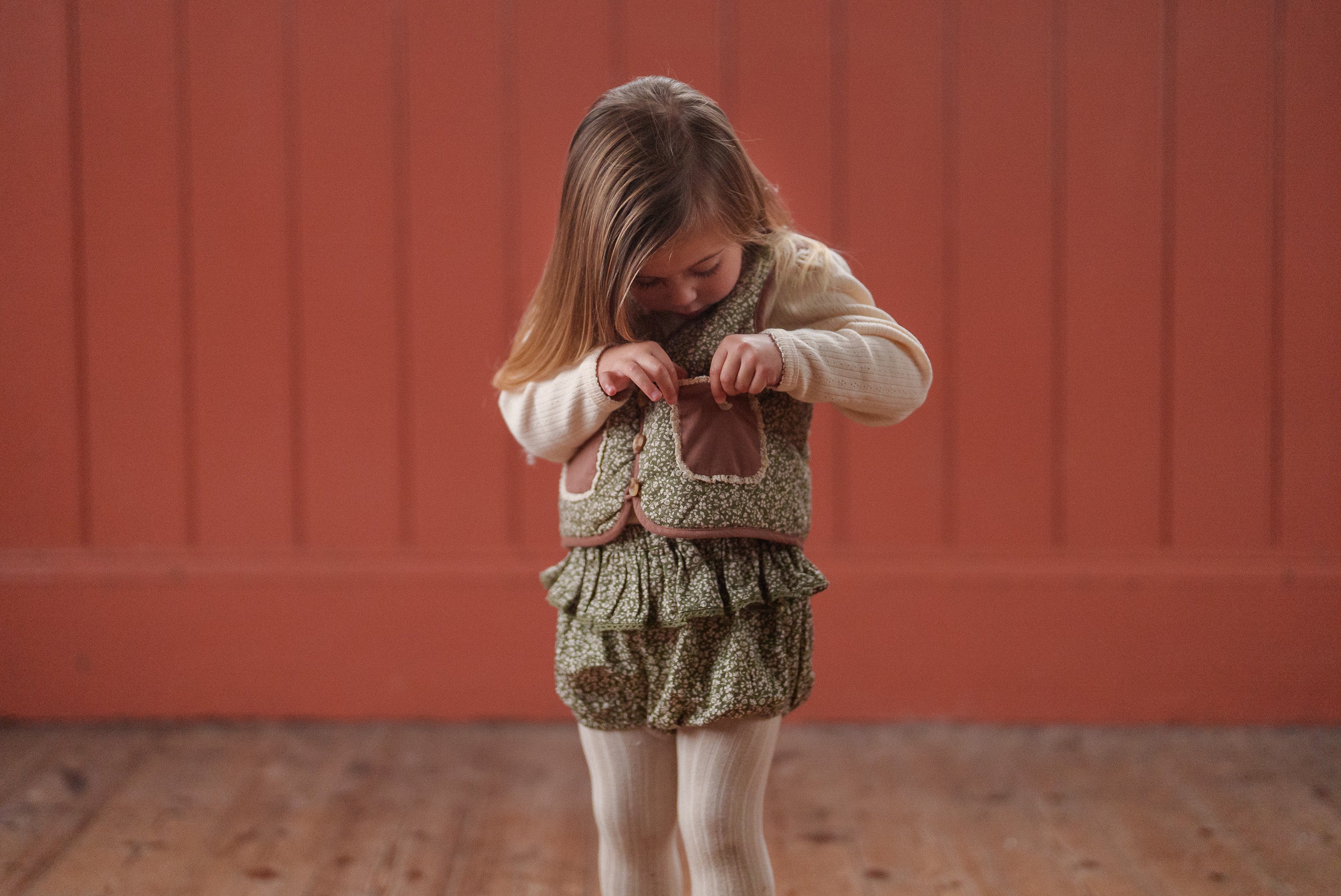 Young girl wearing a green floral organic cotton romper with ruffles and a soft knitted vest, styled in vintage-inspired sustainable children's clothing.