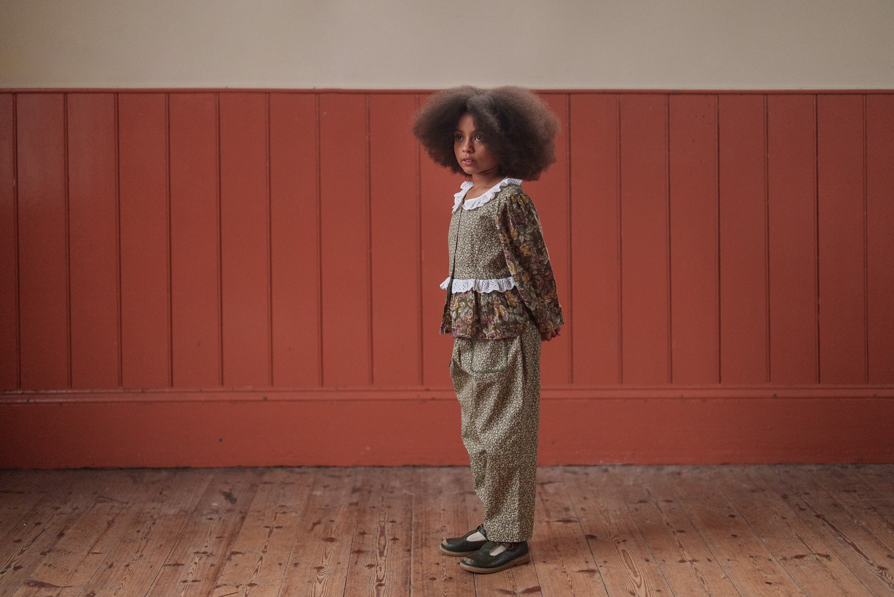 Young girl wearing a vintage style organic cotton floral blouse and matching trousers in soft green and pink, standing indoors against a warm wooden panel backdrop.