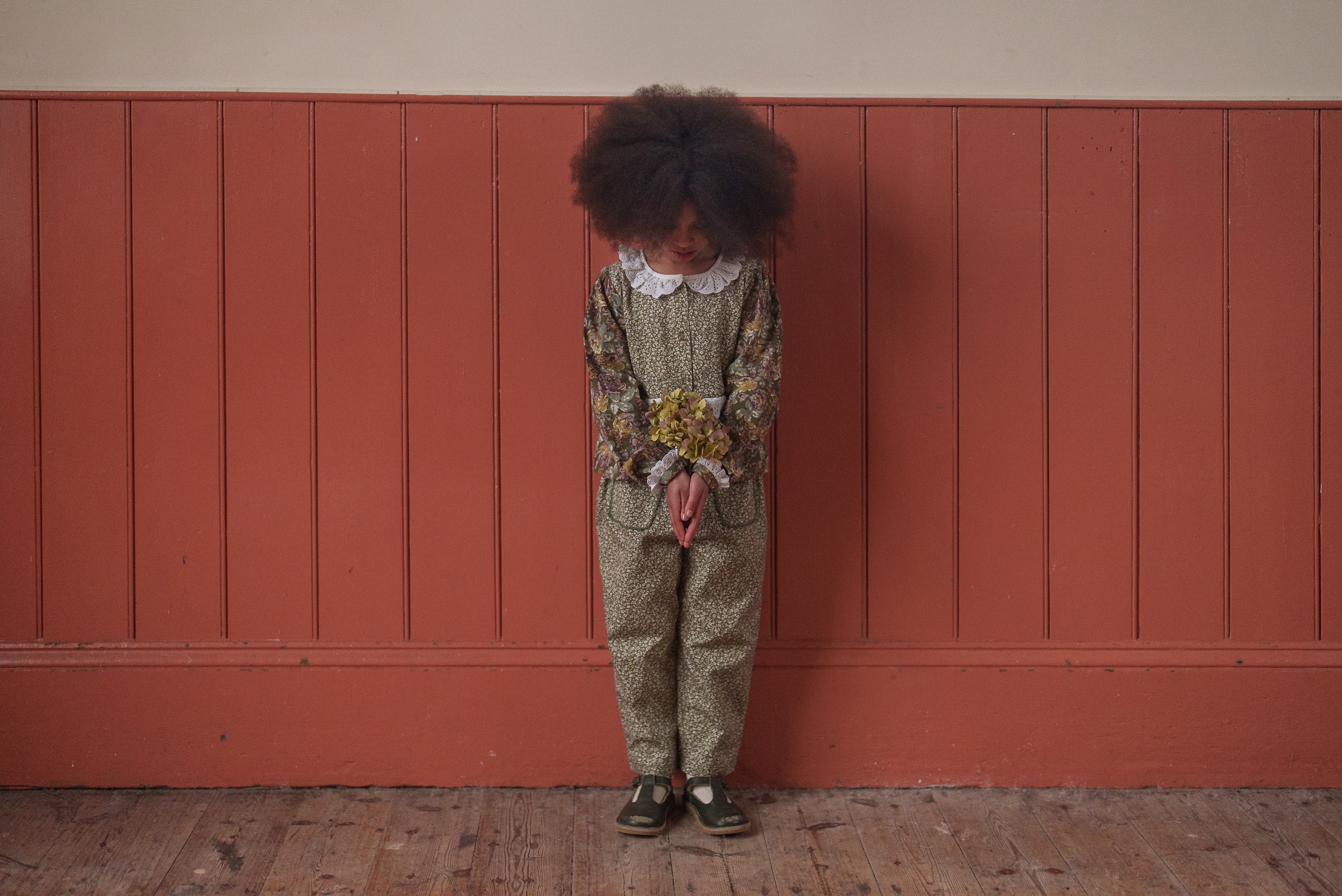 Child wearing a vintage style organic cotton blouse with green and pink floral pattern, holding a small bouquet, standing against a warm terracotta wall.