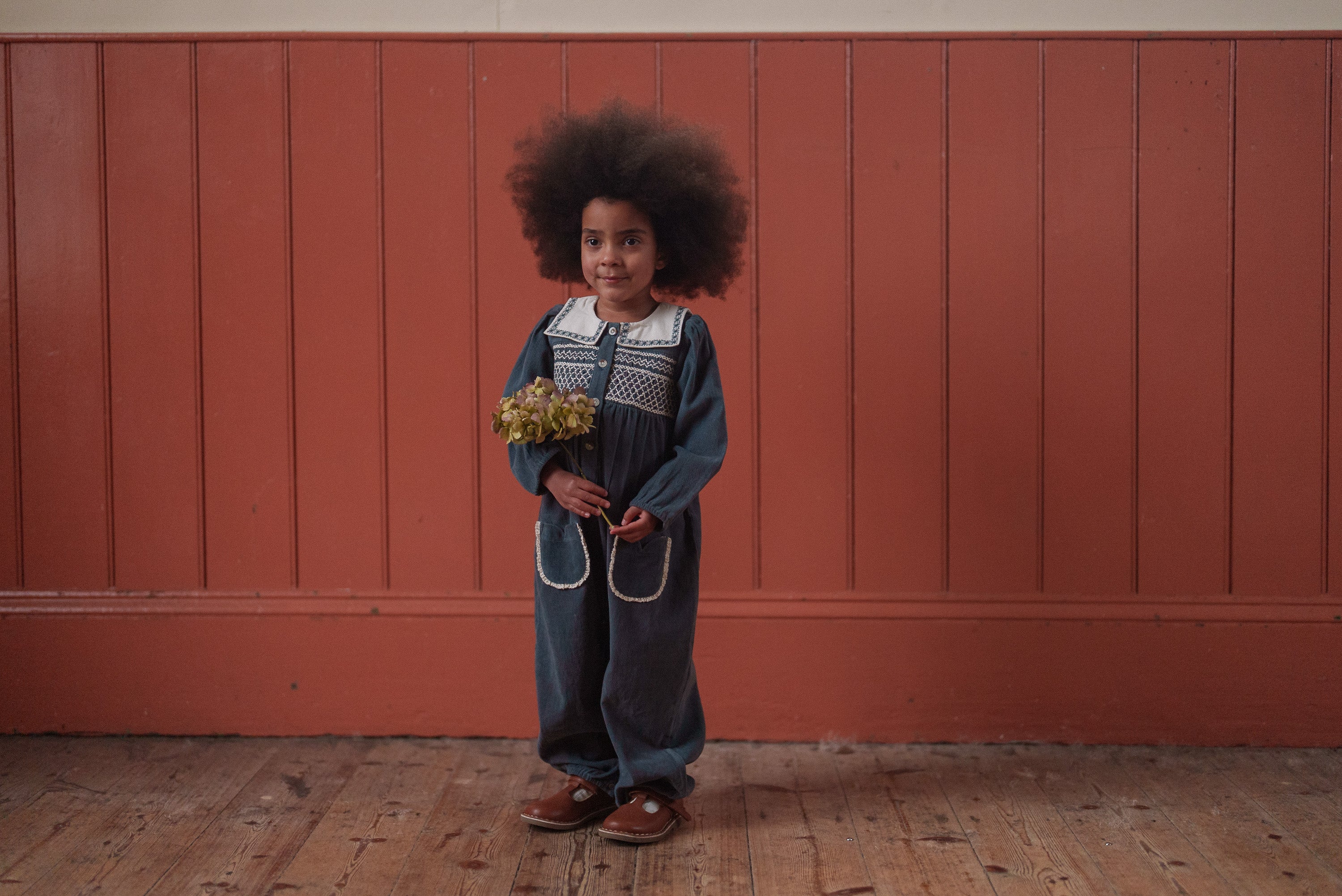 Child wearing a vintage style organic cotton romper in deep green, holding a small bouquet, standing against a rustic wooden wall in natural light.