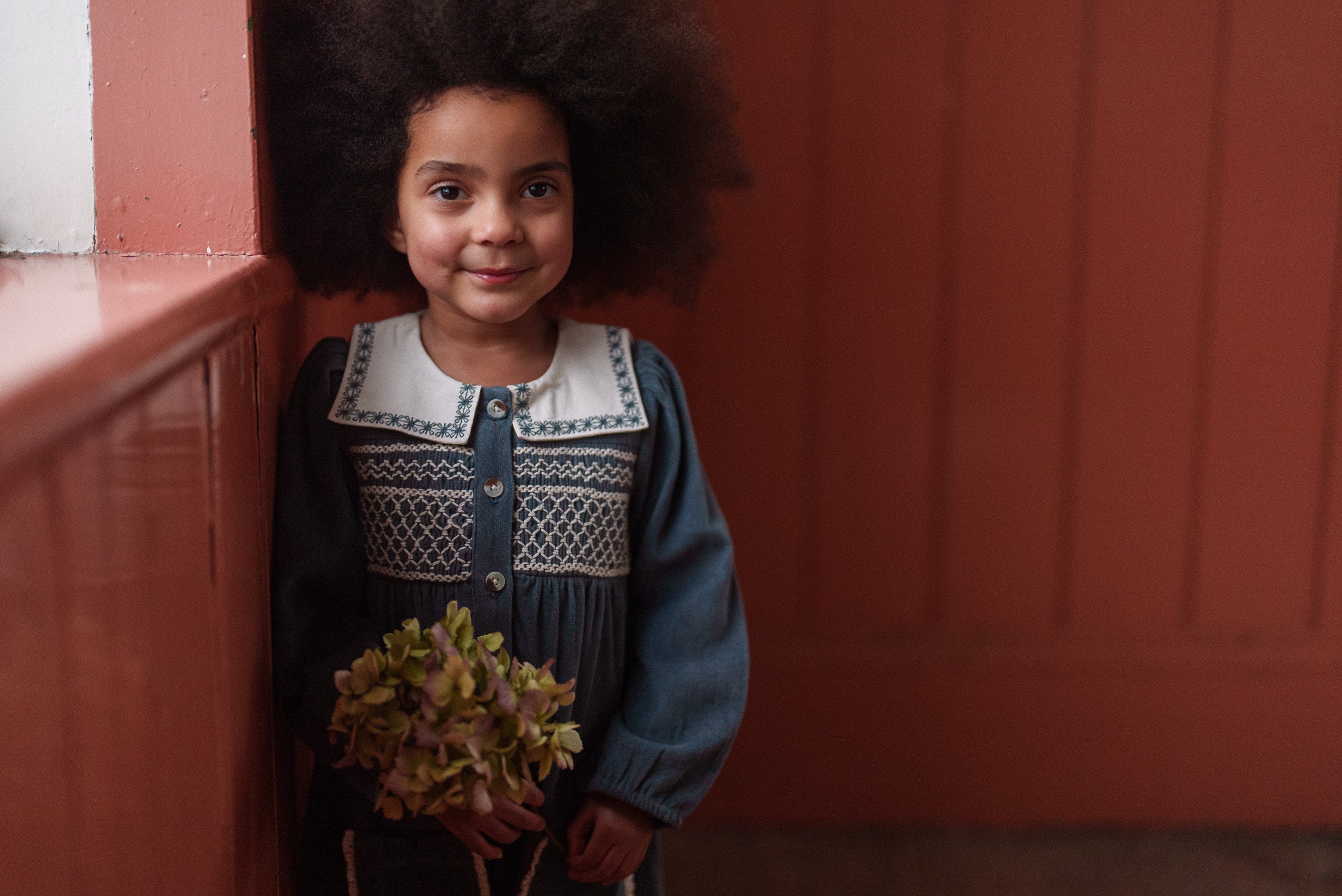 Young girl with natural curly hair wearing a vintage style organic cotton romper in Juniper velvet, holding flowers and standing against a warm-toned wooden wall.