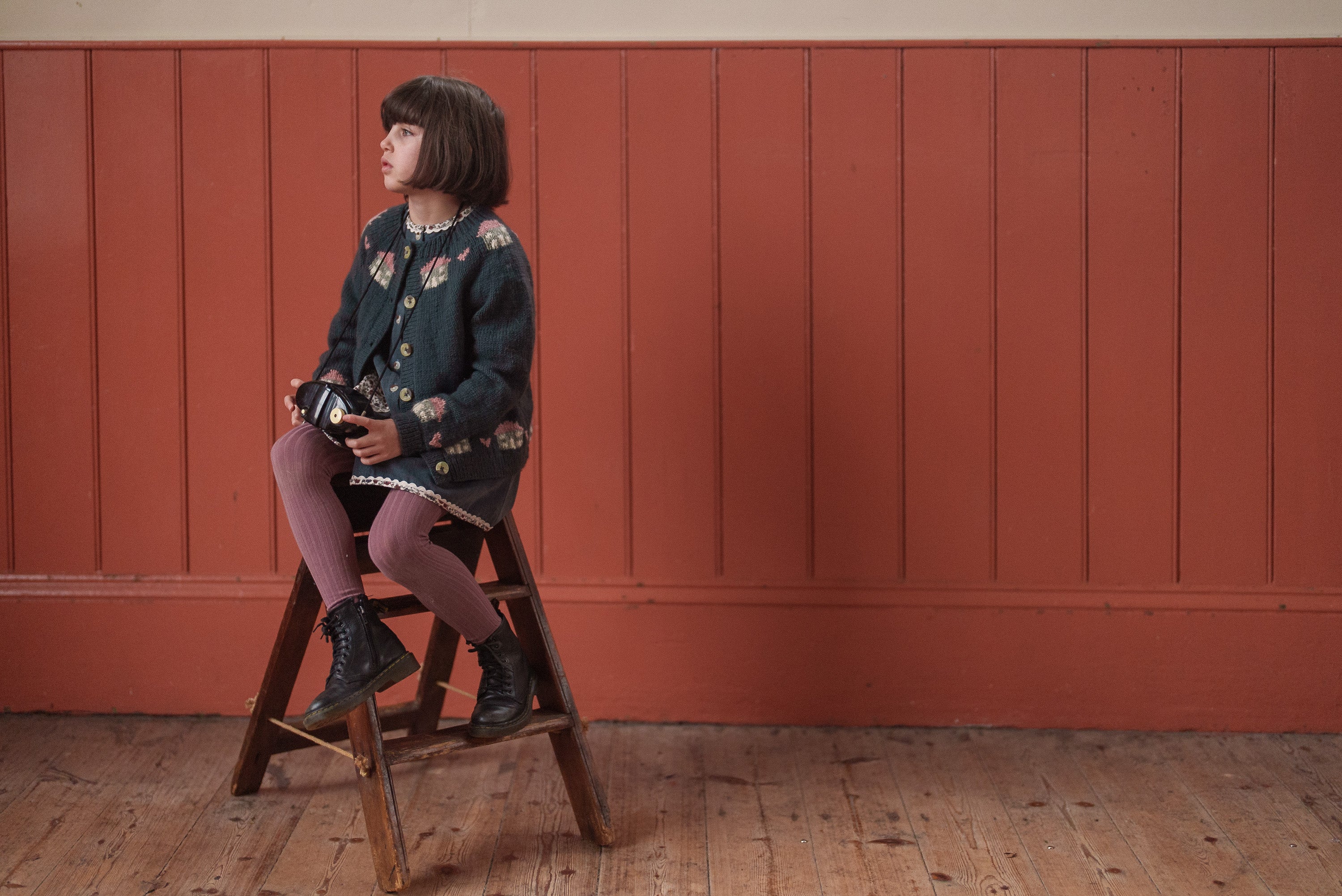 Young child wearing a multicolor, blue hand-knitted merino cardigan with floral details, sitting on a wooden stool against a warm vintage-style wall.
