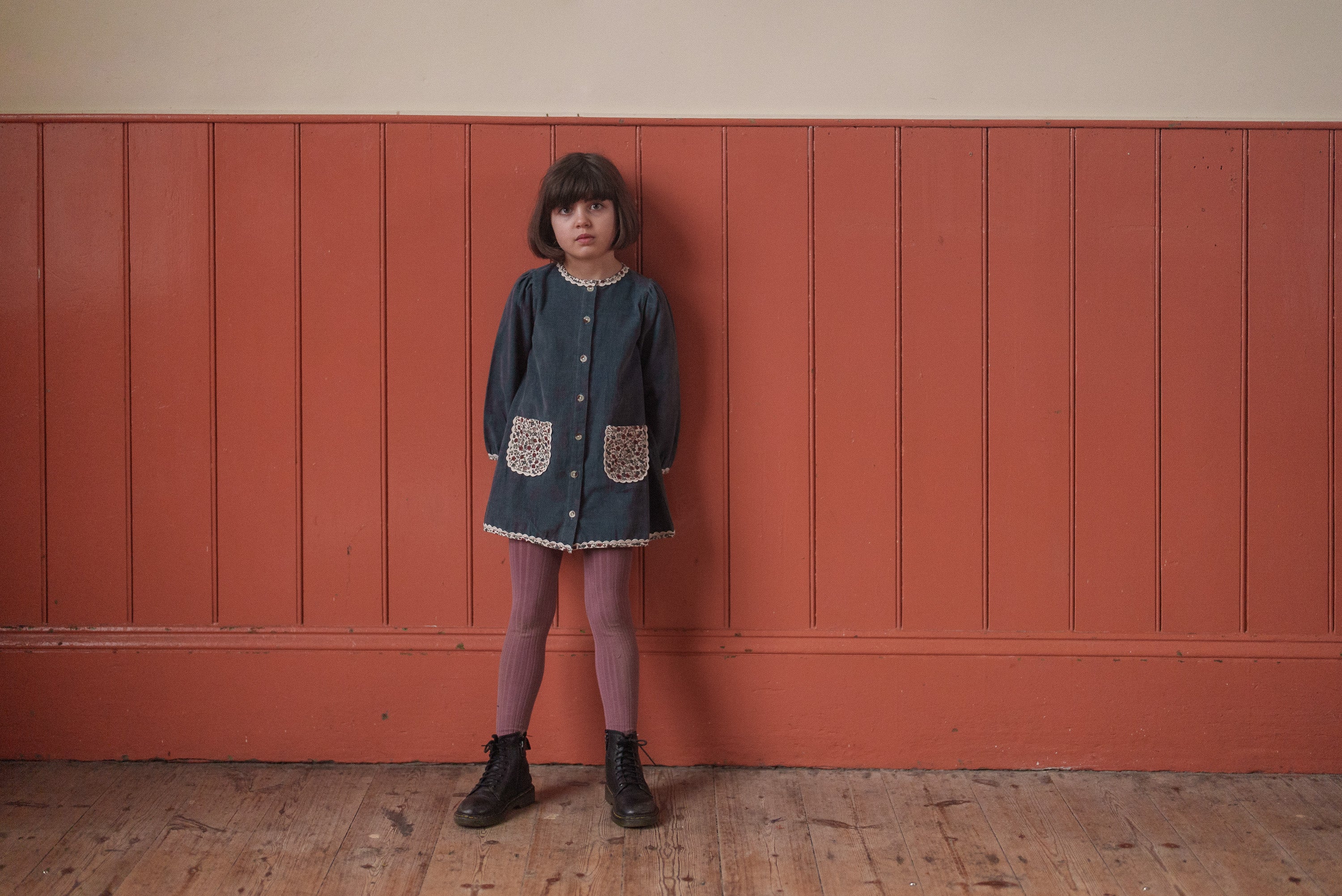Young girl wearing a vintage-style blue velvet dress with floral pockets, organic cotton tights, and black boots, standing indoors against a rustic wood panel background.