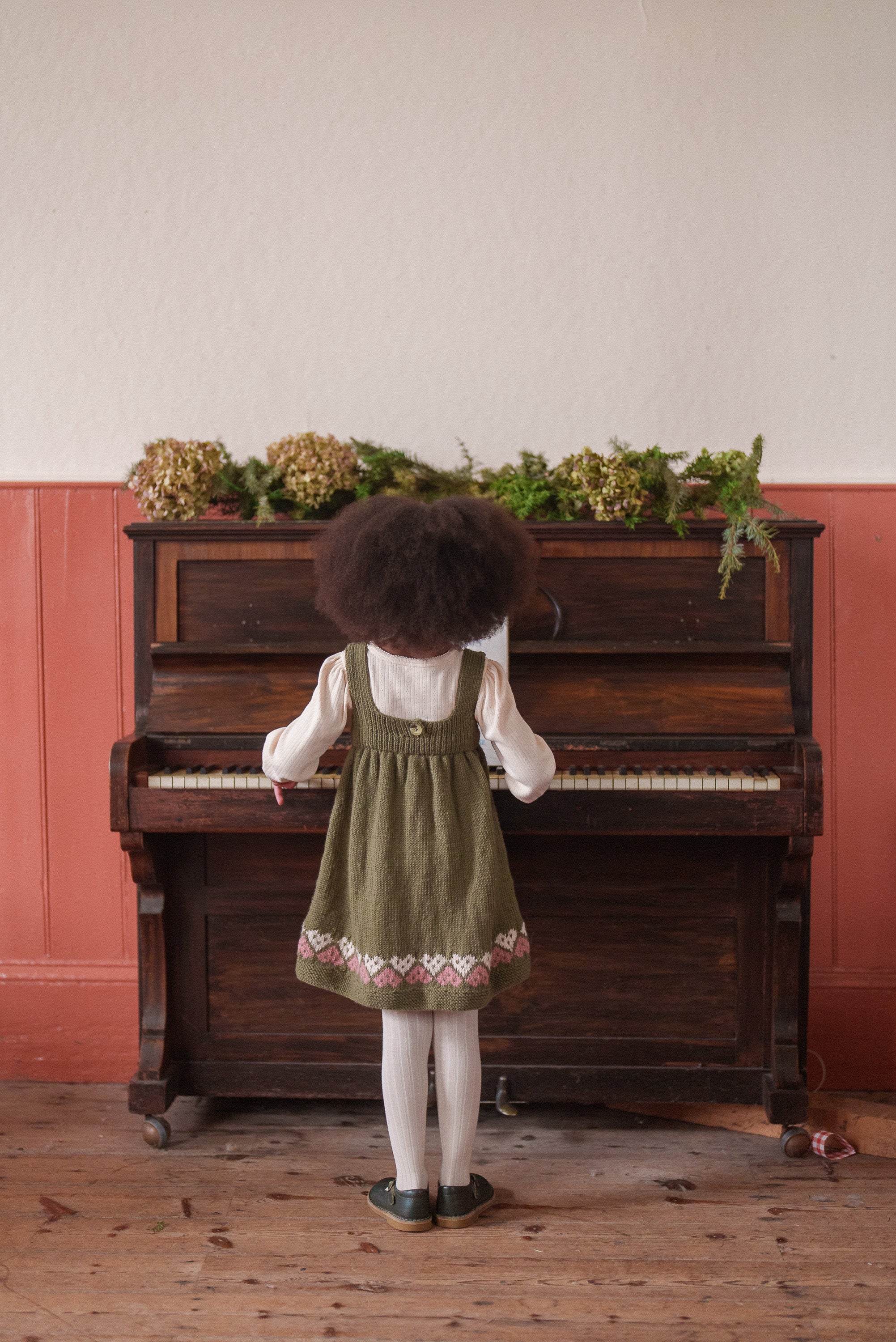 Child wearing a hand-knitted pinafore dress in olive green with heart patterns, playing a vintage wooden piano in a cozy room with natural decor.