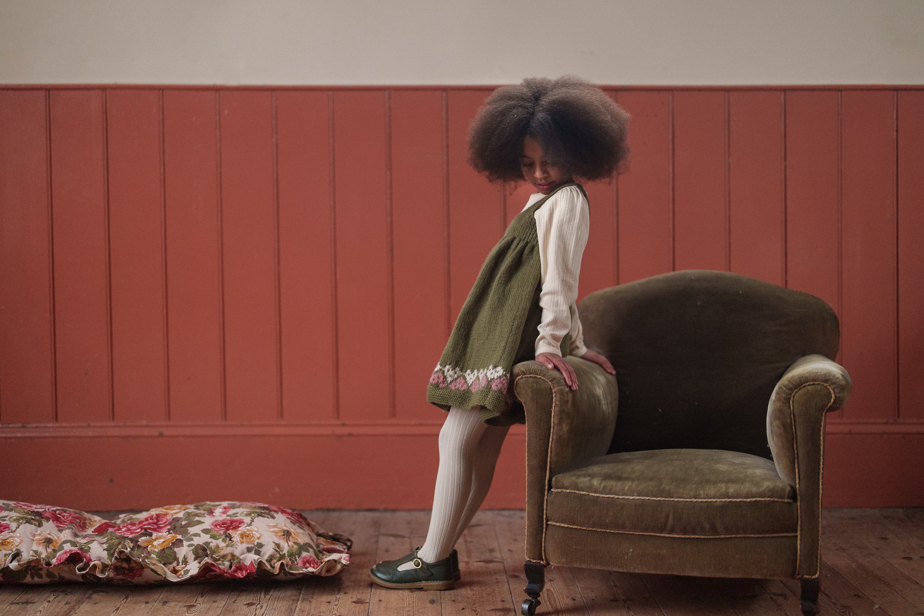 Child wearing a navy and beige hand-knitted pinafore dress with heart details, standing beside a vintage green armchair in a warm, natural light setting