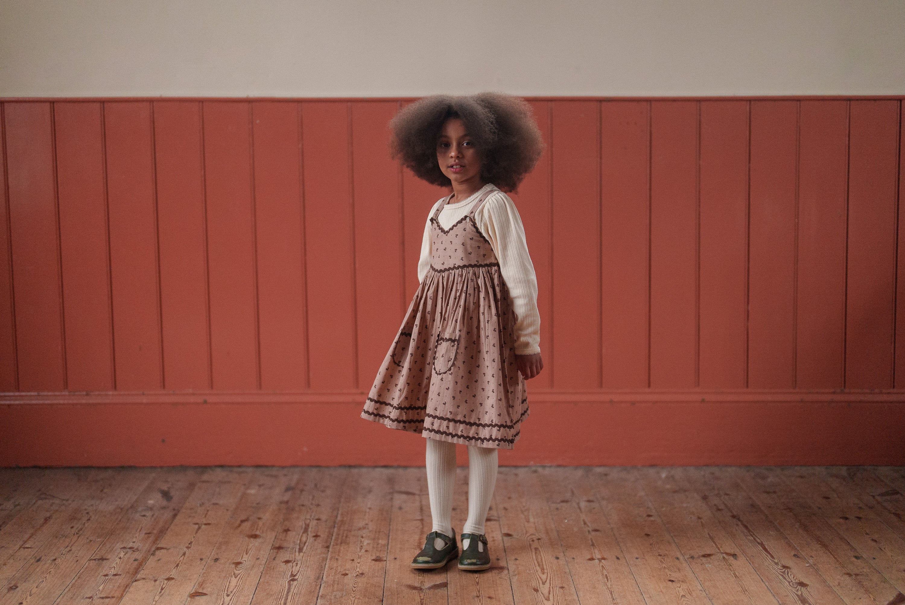 Young girl wearing a vintage style dress over a cream organic cotton stretchable T-shirt, standing on wooden floor against a warm toned wall, showcasing sustainable children's clothing.