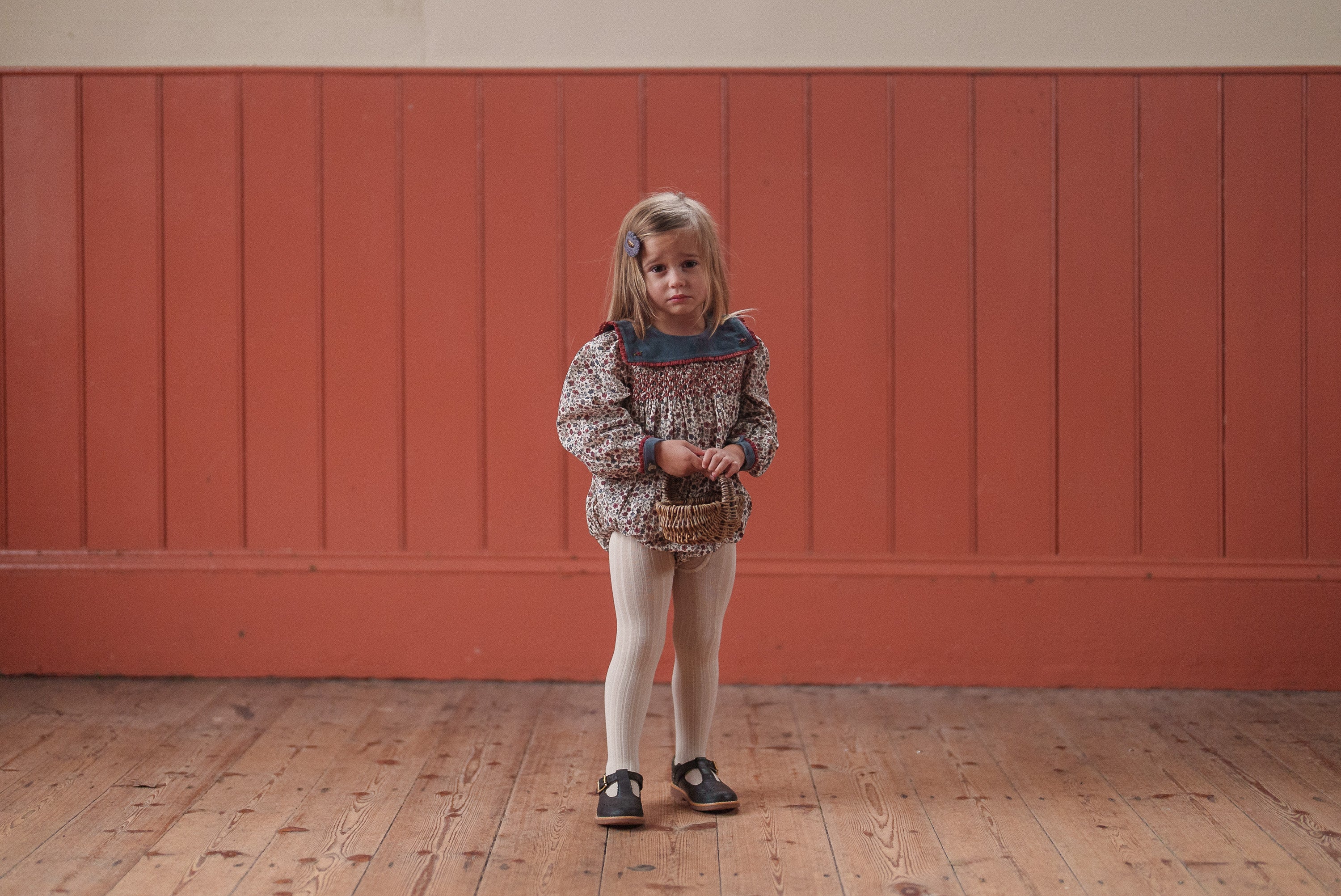 Young girl wearing vintage-style organic cotton romper with floral print and tights, holding a small wicker basket, standing on wooden floor against warm-toned wall.