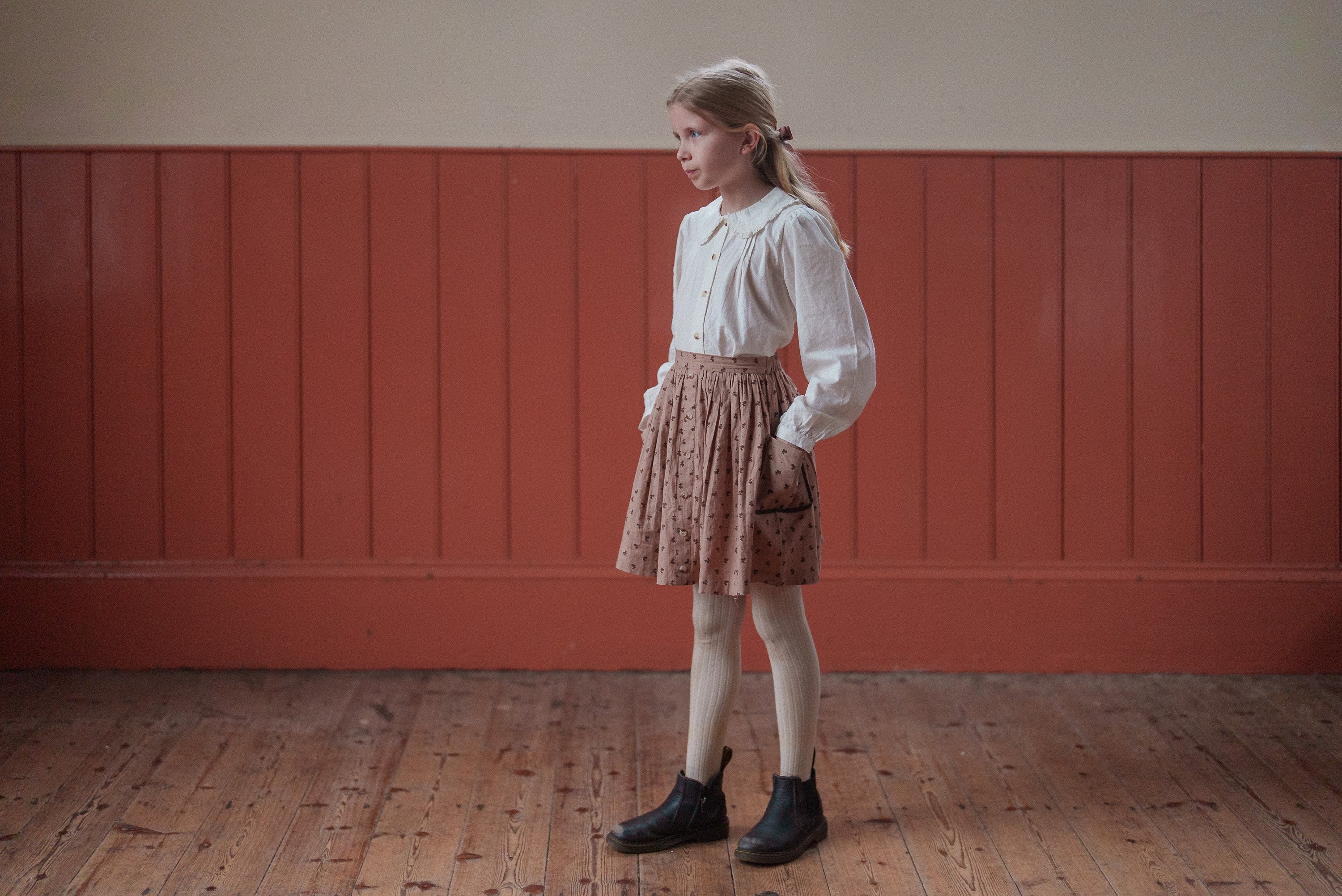 Young girl wearing a vintage-style skirt with a rosehip floral pattern made from organic cotton, paired with a classic white blouse and boots, standing indoors.