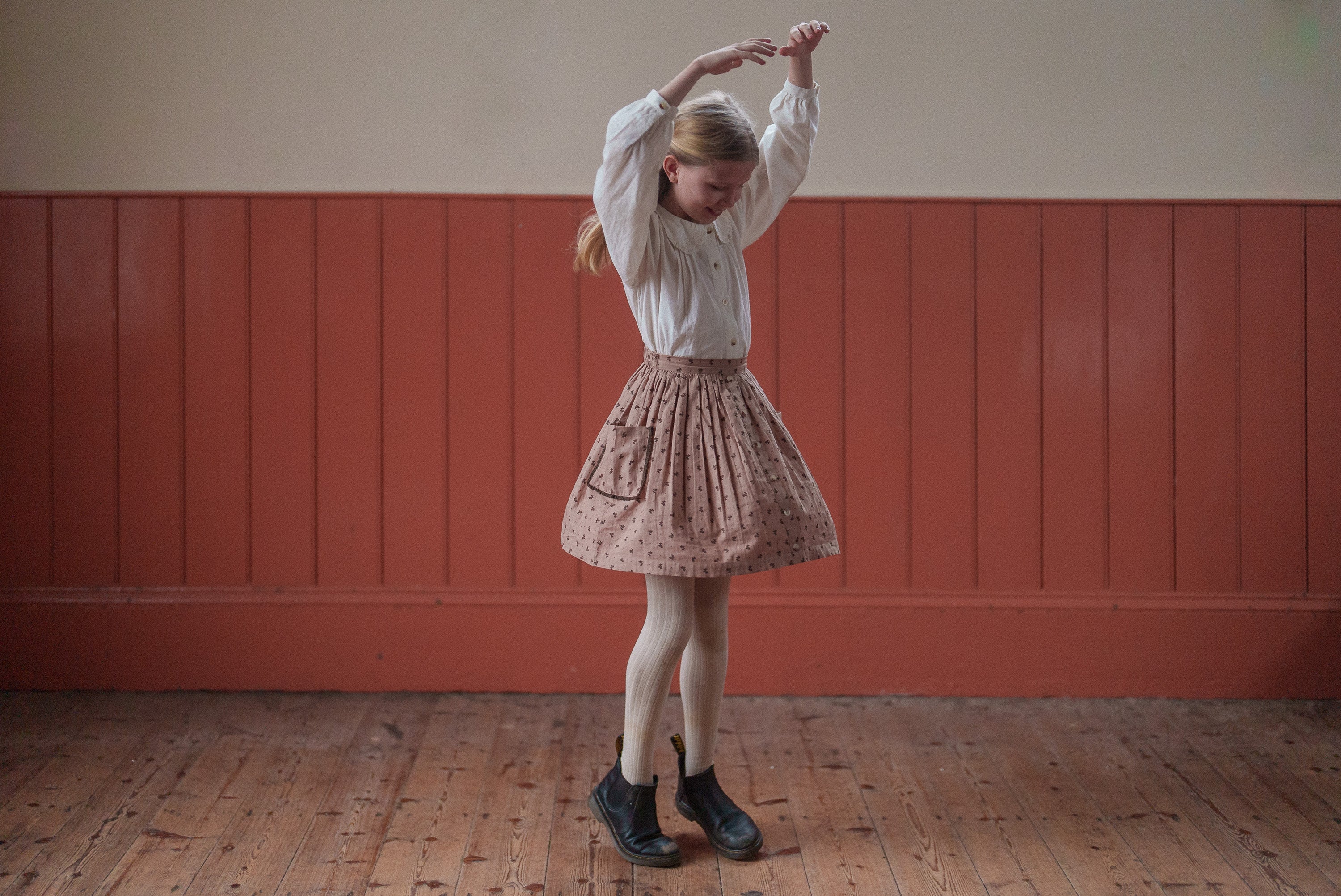 Young girl wearing a vintage-inspired organic cotton pink floral skirt paired with a white blouse, showcasing sustainable, ethically made children’s clothing in a warm, natural setting.