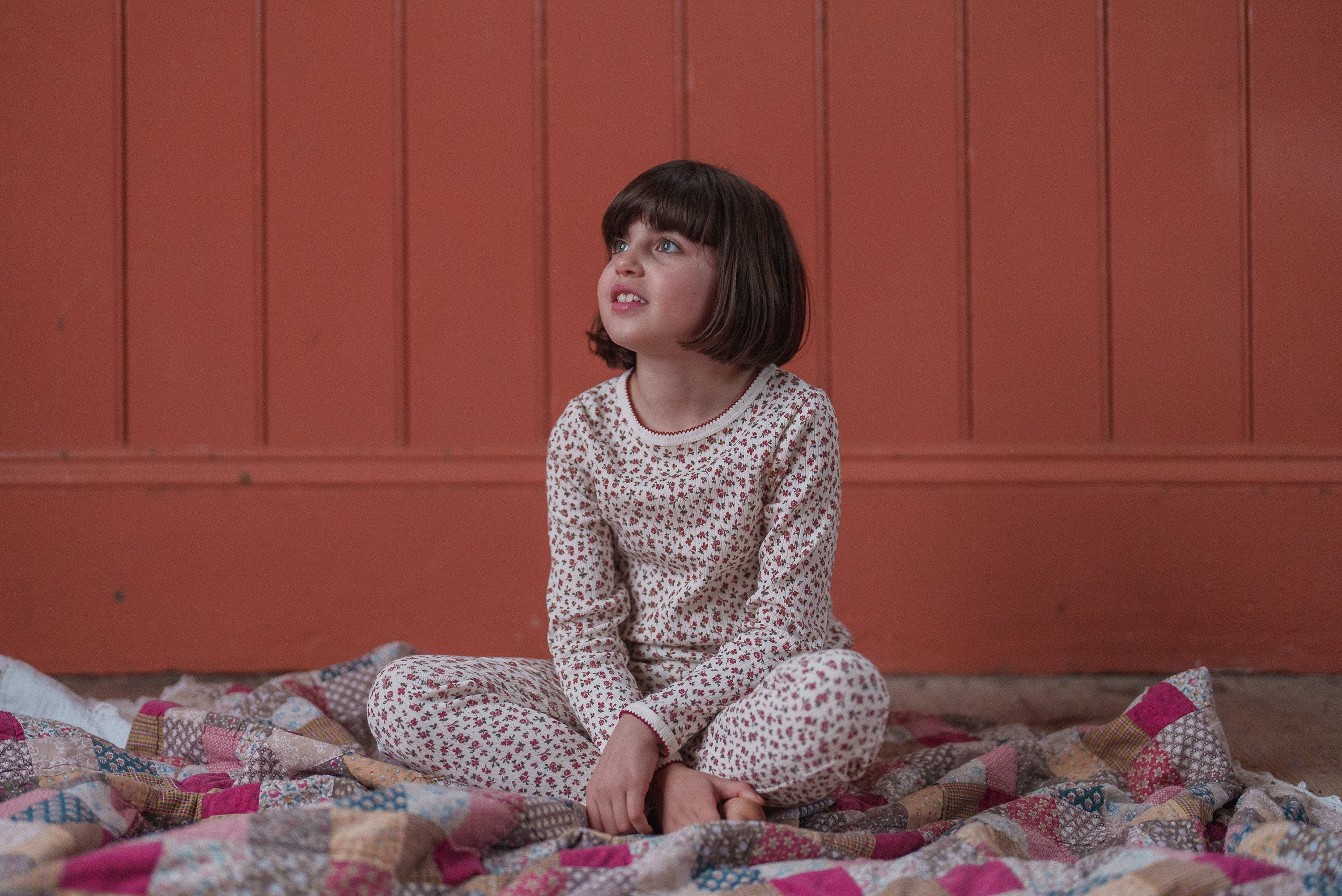 A young child wearing organic jersey cotton pyjamas with a delicate floral pattern, sitting comfortably on a patchwork quilt against a warm, vintage-style wooden backdrop.