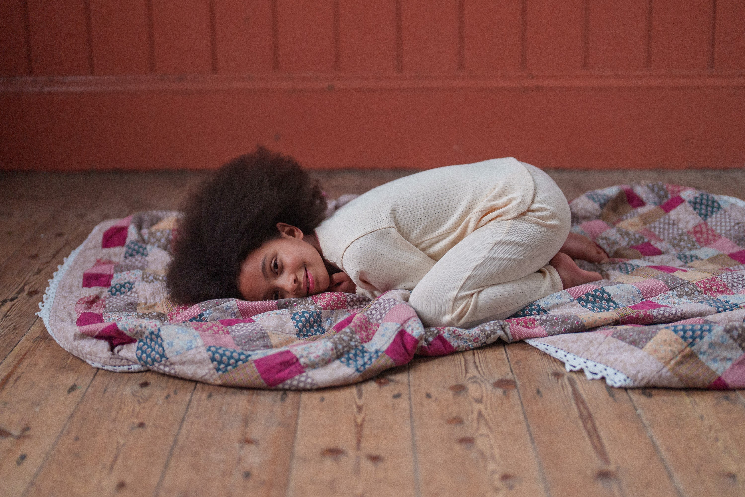Young child wearing soft buttermilk-colored pyjamas lying curled up on a vintage patchwork quilt on wooden floor, showcasing natural and ethical children's clothing.
