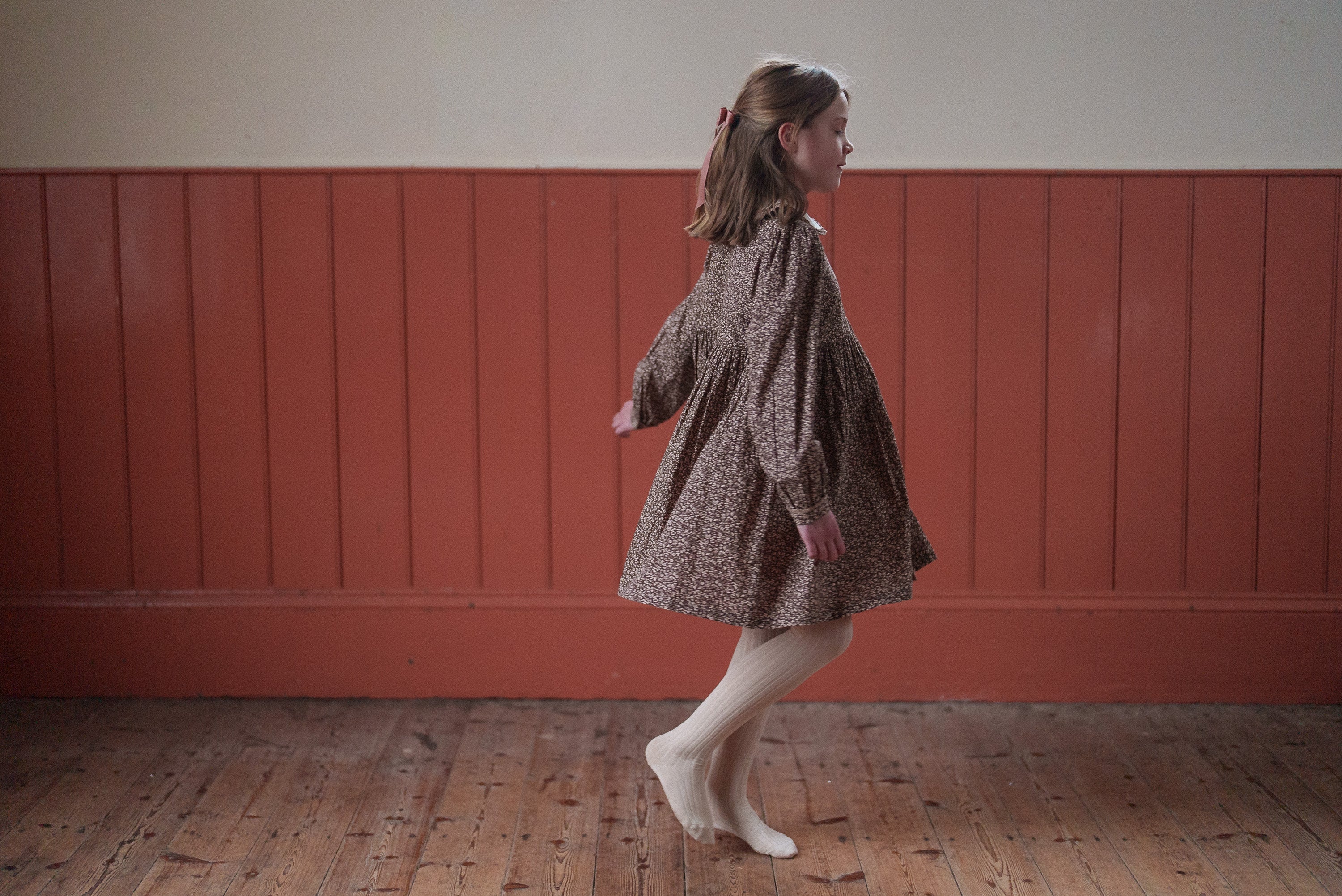 Young girl twirling indoors wearing a brown and beige floral organic cotton dress with vintage style, paired with cream tights in a warm, natural setting.