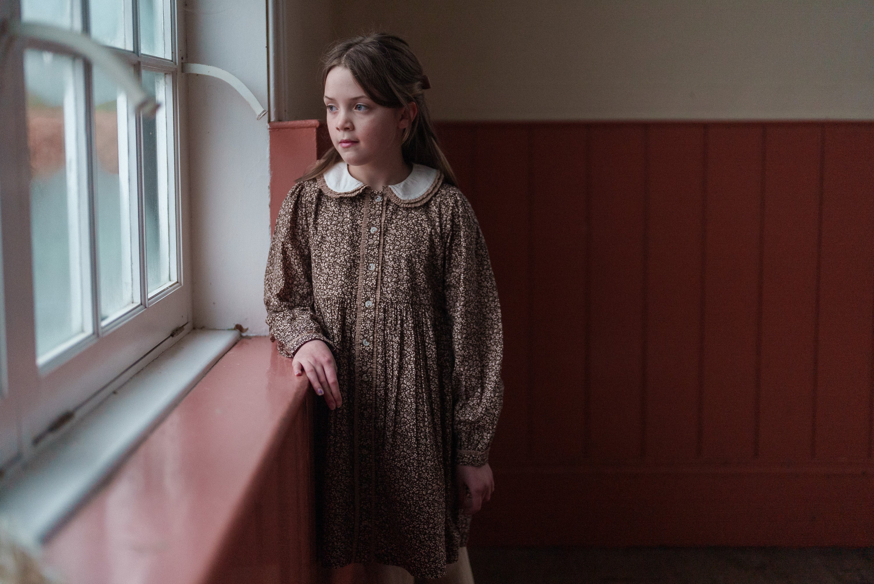 Young girl wearing a brown and beige floral organic cotton dress with a vintage style, standing by a window in a softly lit room, showcasing sustainable children’s clothing.