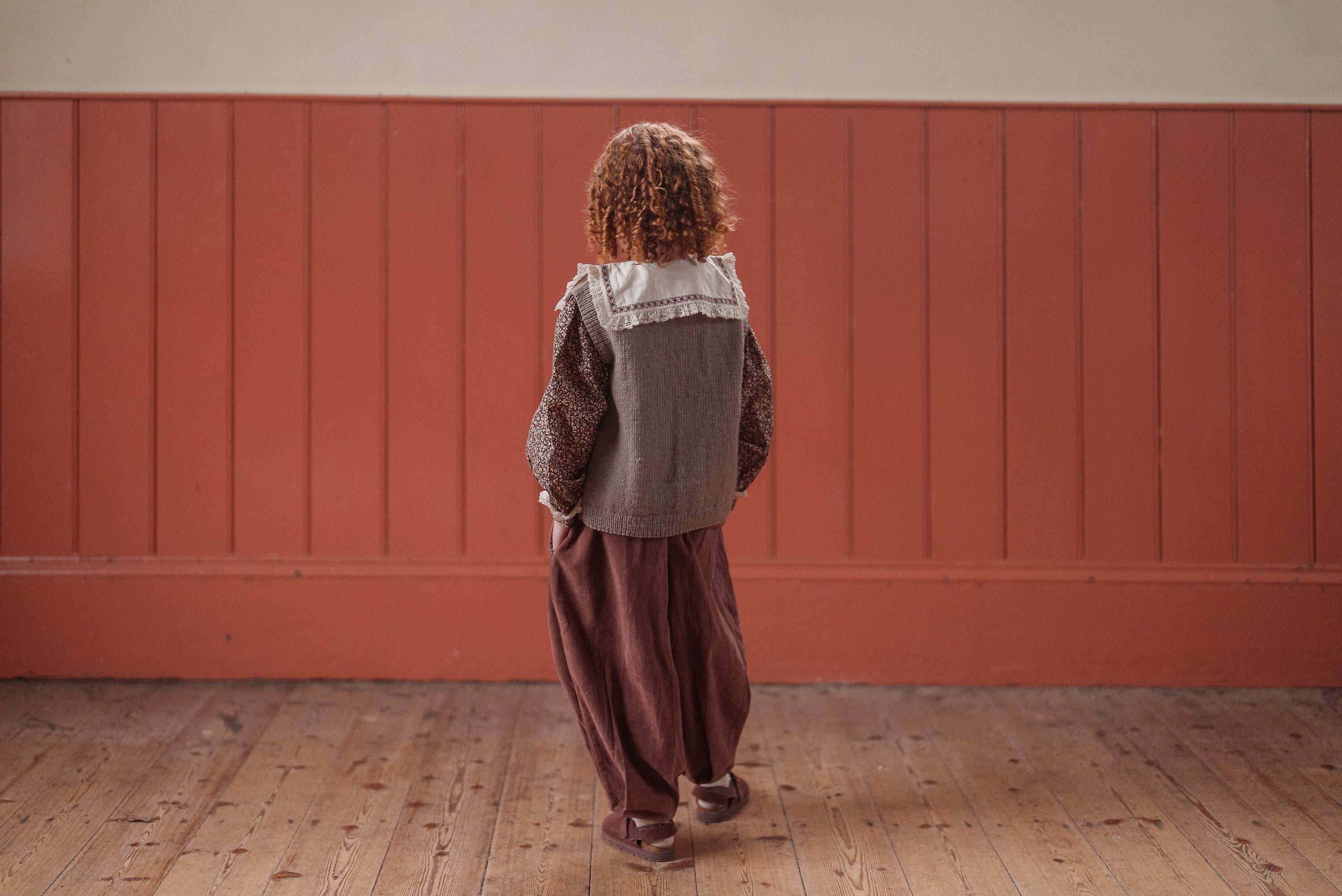 Child wearing vintage style sustainable children's clothing in earthy tones, featuring a hand-knitted vest made of natural merino wool, standing on a wooden floor.