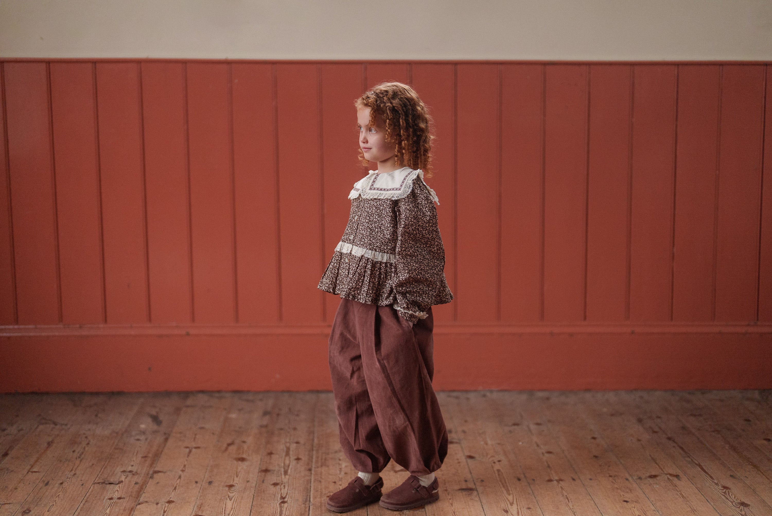 Child wearing vintage style children's clothing in warm brown tones, featuring natural fabrics and a relaxed fit, standing on wooden floor against rustic wall.