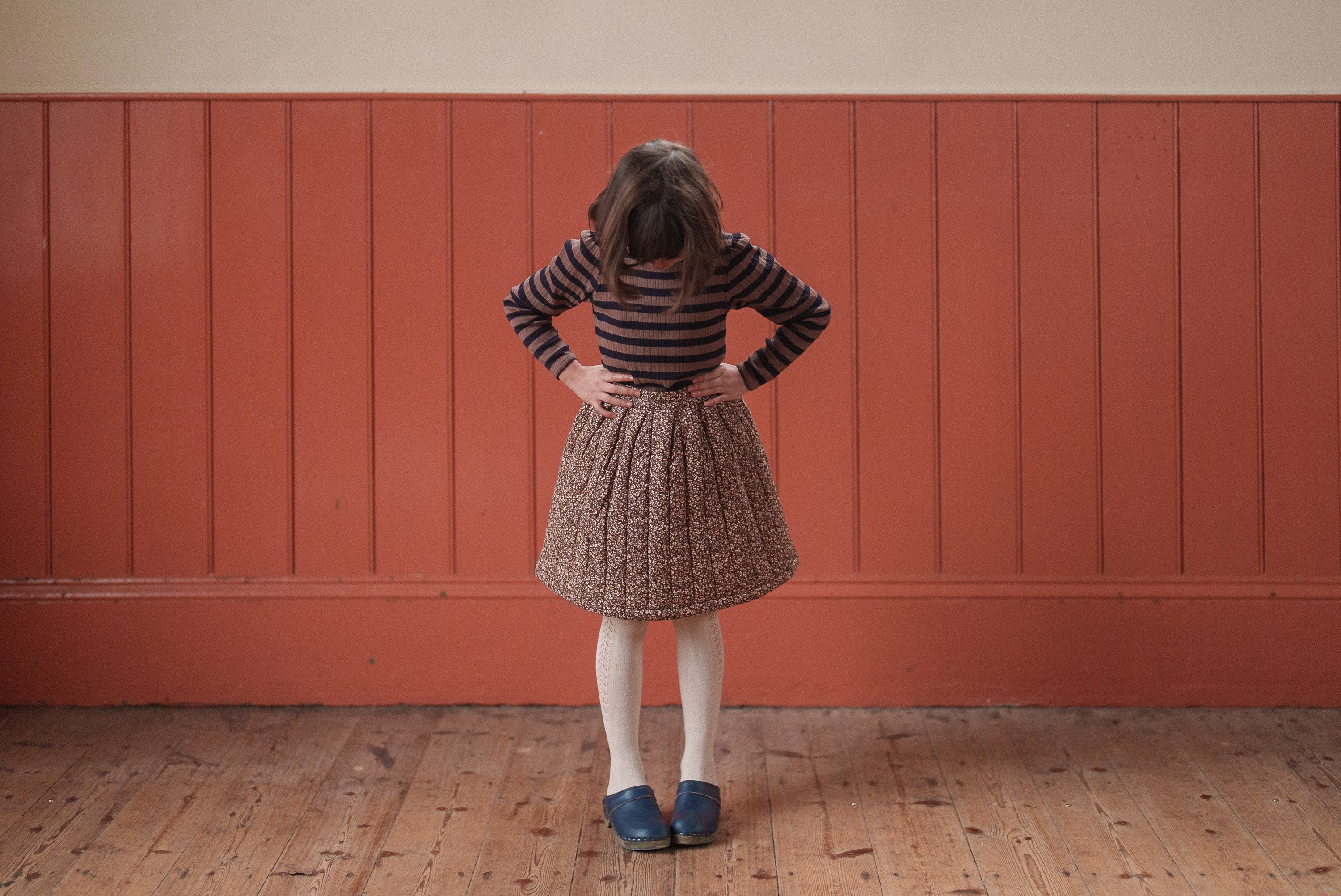 Child wearing navy and beige striped slim fit organic cotton t-shirt with vintage style skirt and navy shoes, standing on wooden floor against red paneled wall.