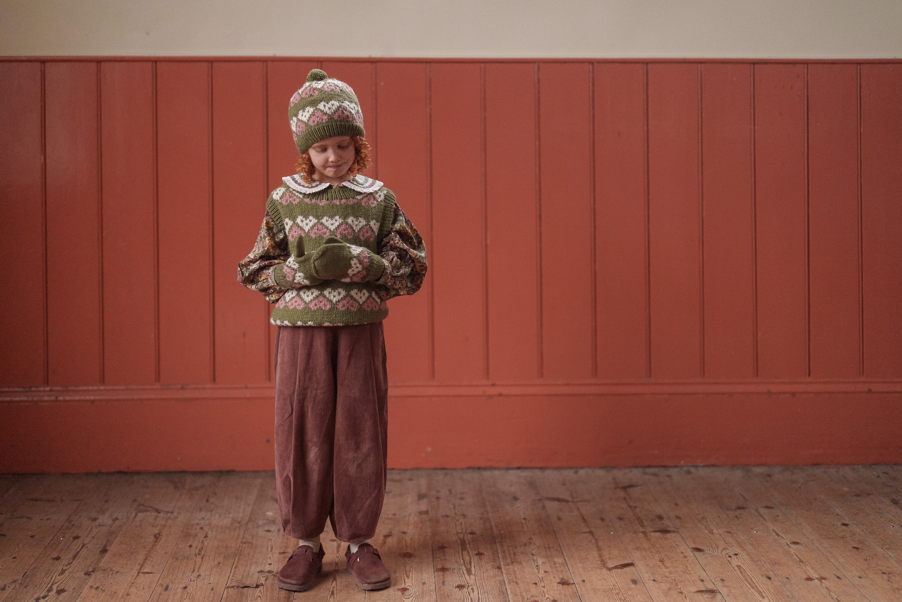Child wearing vintage style children's clothing in earthy tones, including a knit sweater, matching hat and mittens, and loose brown trousers on wooden floor with warm toned wall background.