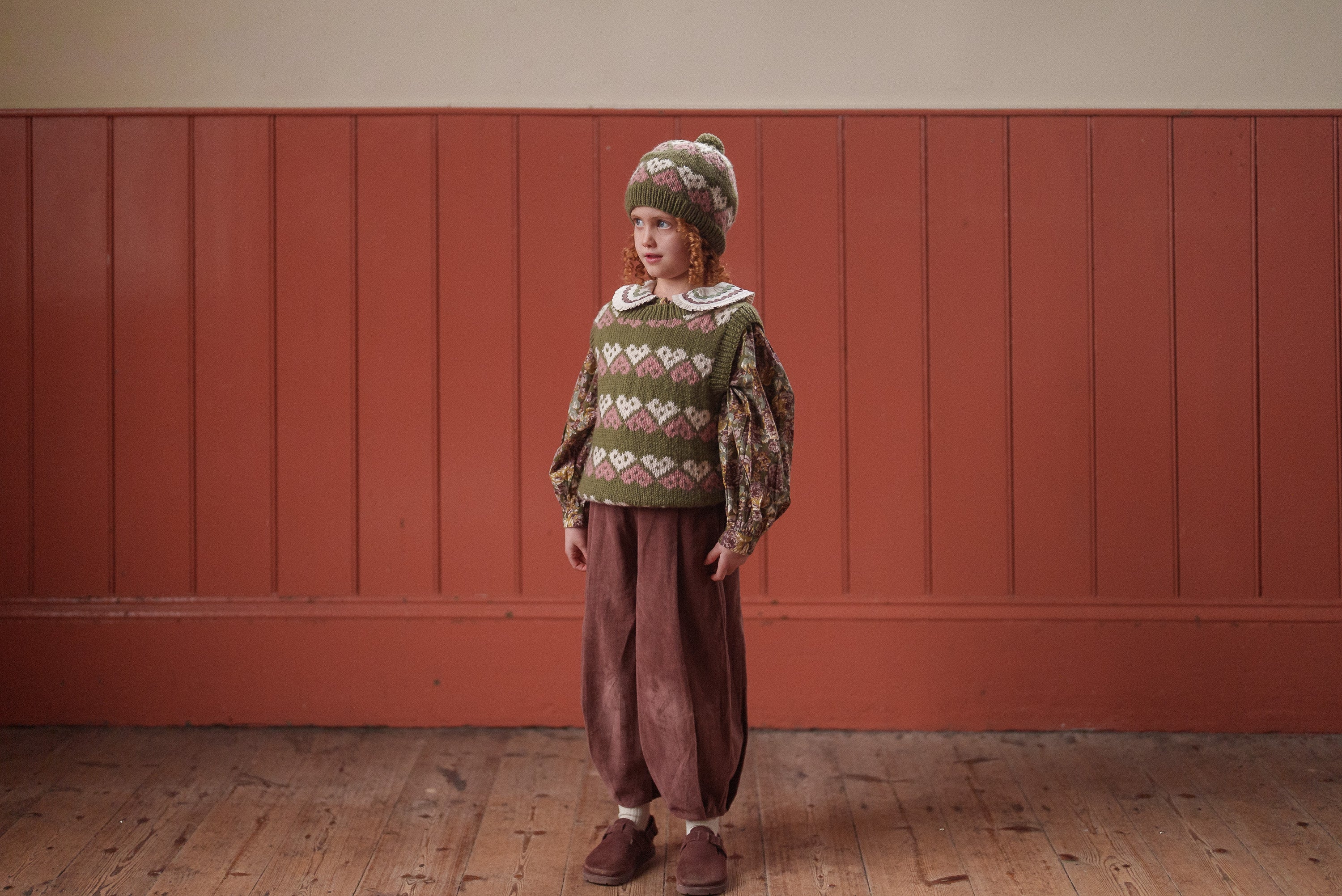 Child wearing vintage-style children’s clothing with a multicolor hand-knitted hearts woolen hat and vest, styled in organic, sustainable, ethically made fabrics.