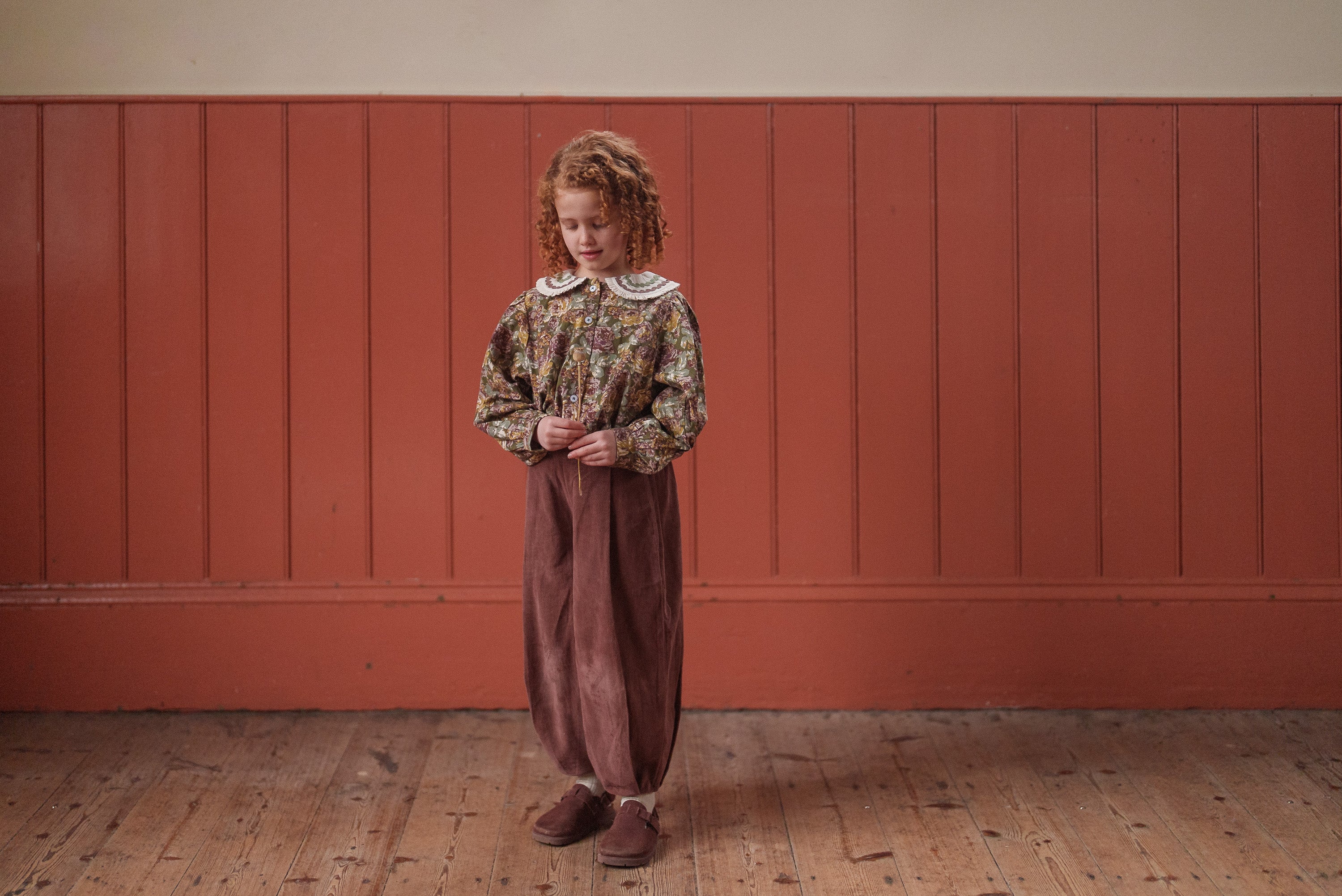 Young girl wearing a vintage style blouse with a floral green and pink pattern made from organic cotton, paired with loose brown pants, standing on a wooden floor against a rustic red wall.