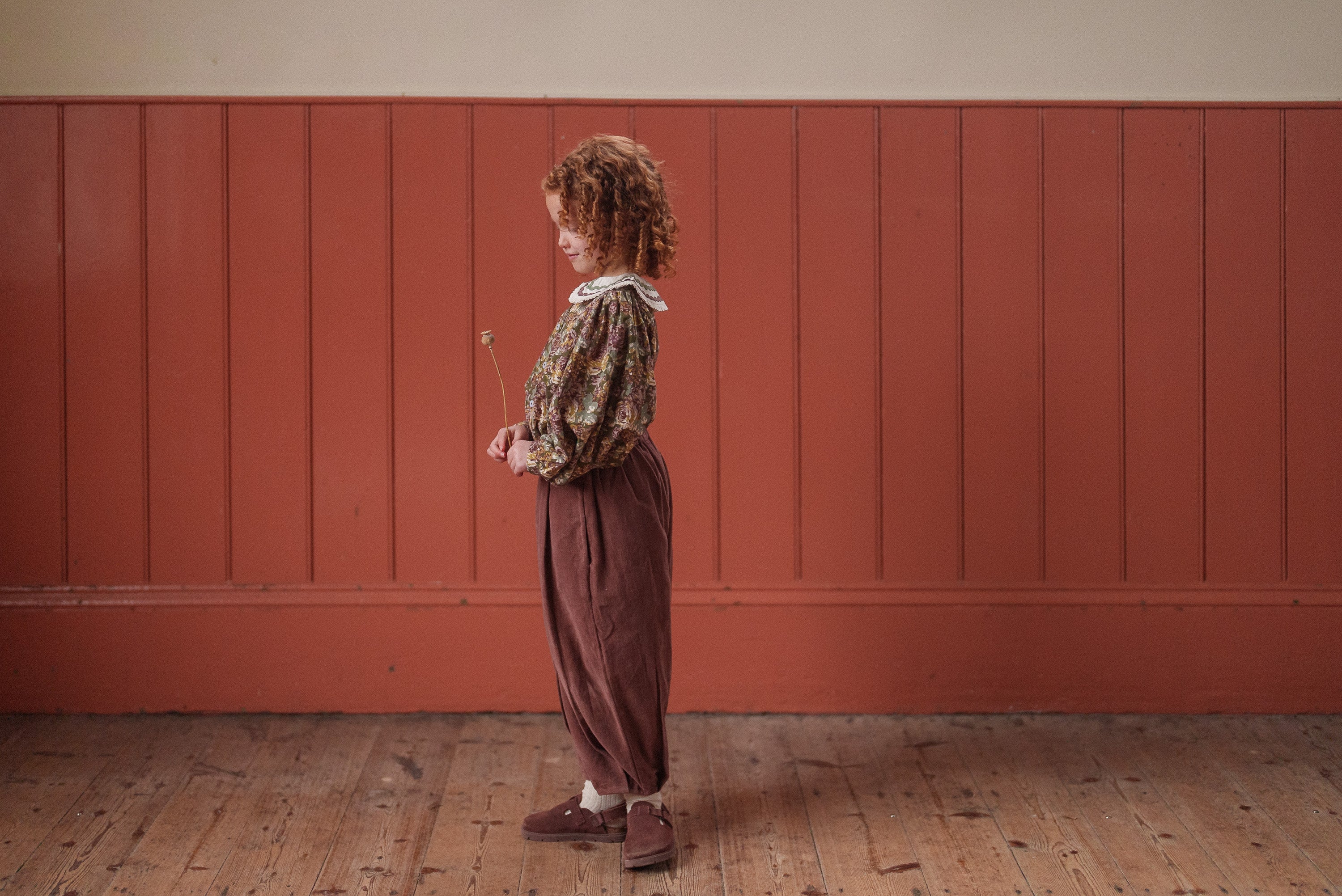 Young child wearing vintage style girls blouse in green and pink floral organic cotton, paired with loose brown trousers, standing indoors on wooden floor against paneled wall.