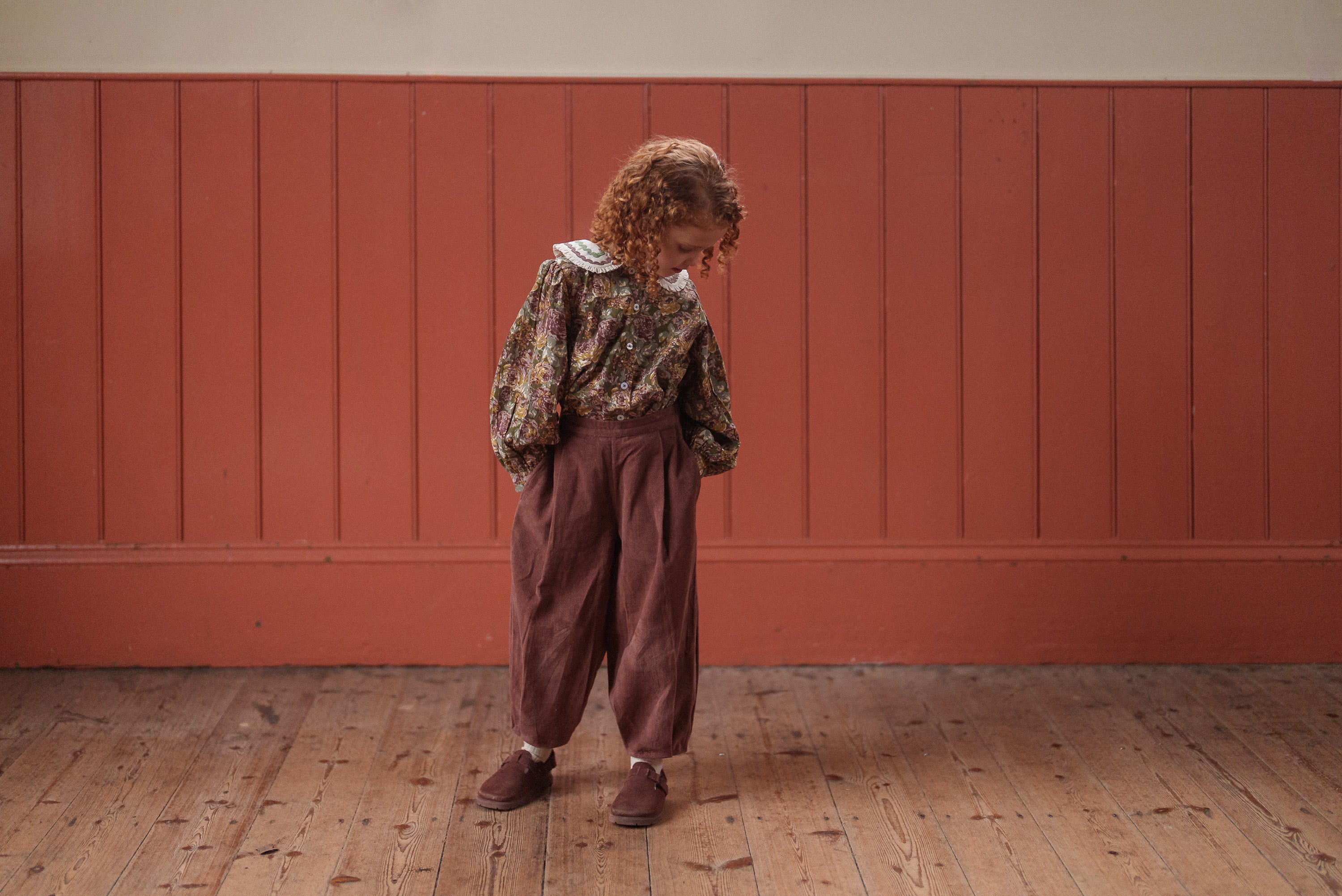 A child wearing a vintage-style girls blouse with green and pink floral print made from organic cotton, paired with loose brown trousers and brown shoes, standing on a wooden floor against a warm-toned wall.