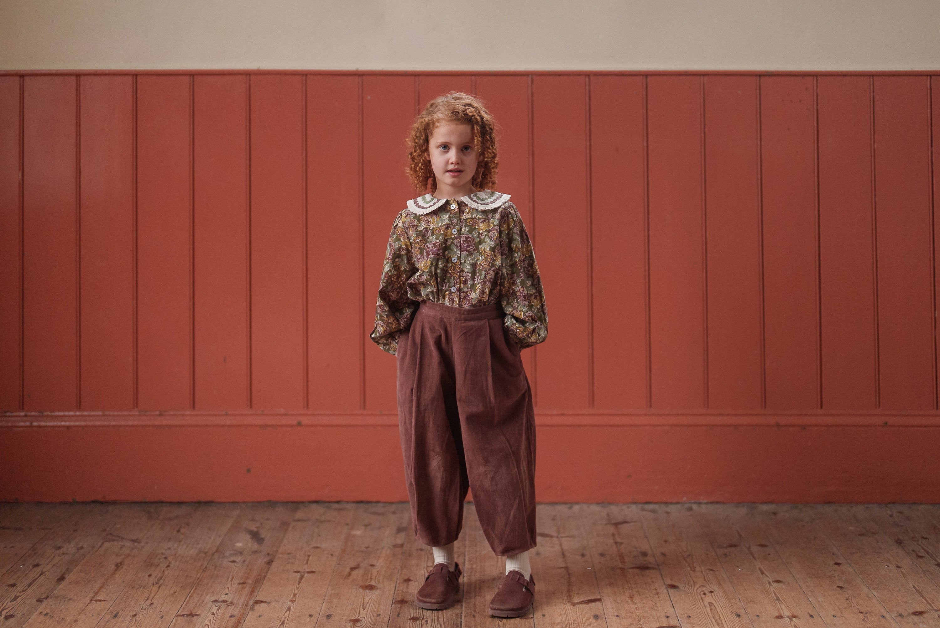 Young child wearing a green and pink floral organic cotton blouse with a vintage style, paired with loose brown trousers and standing against a rustic wooden wall.