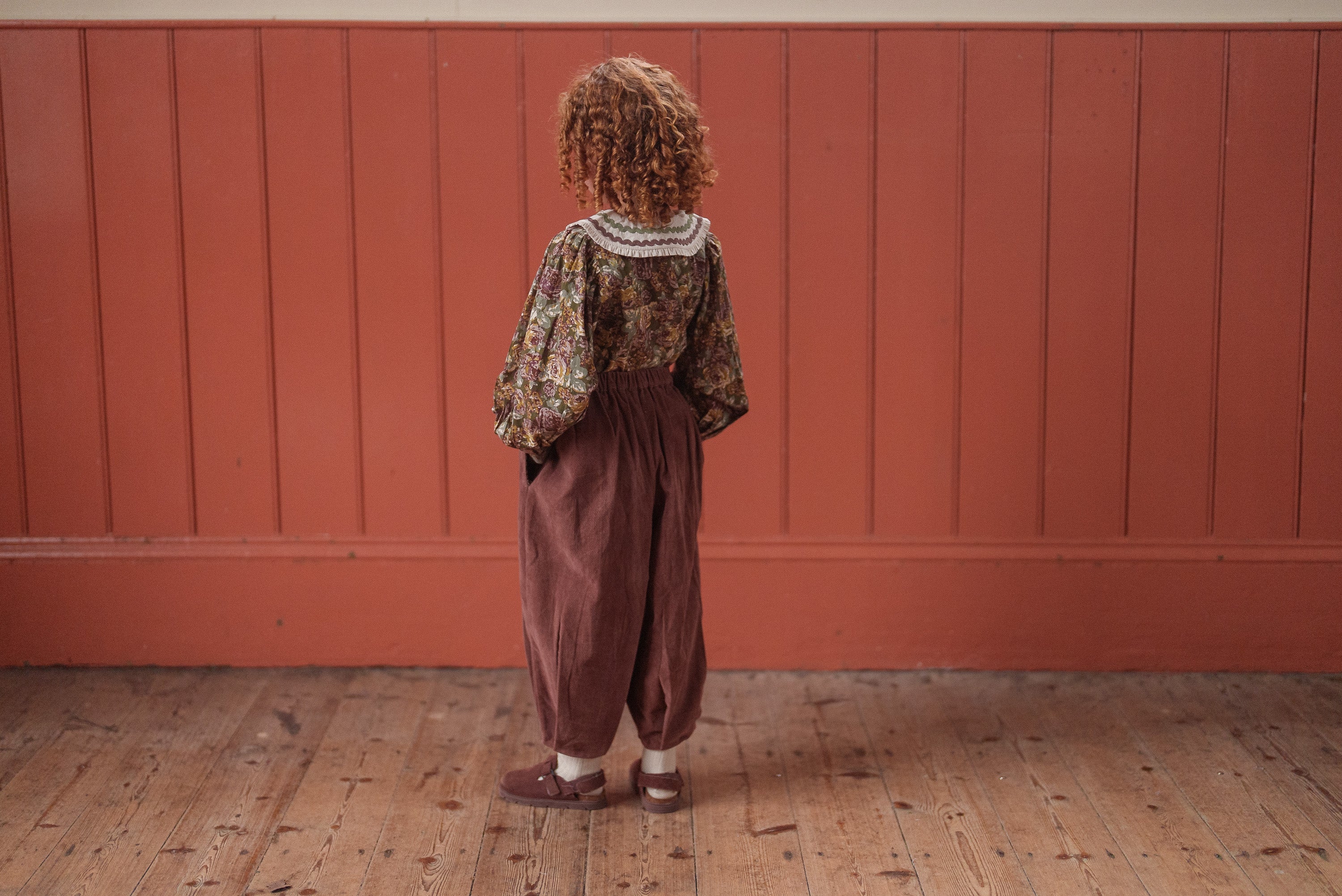 Child wearing vintage-style children's clothing featuring a green and pink floral organic cotton blouse with puffed sleeves and loose brown pants, standing on wooden floor.