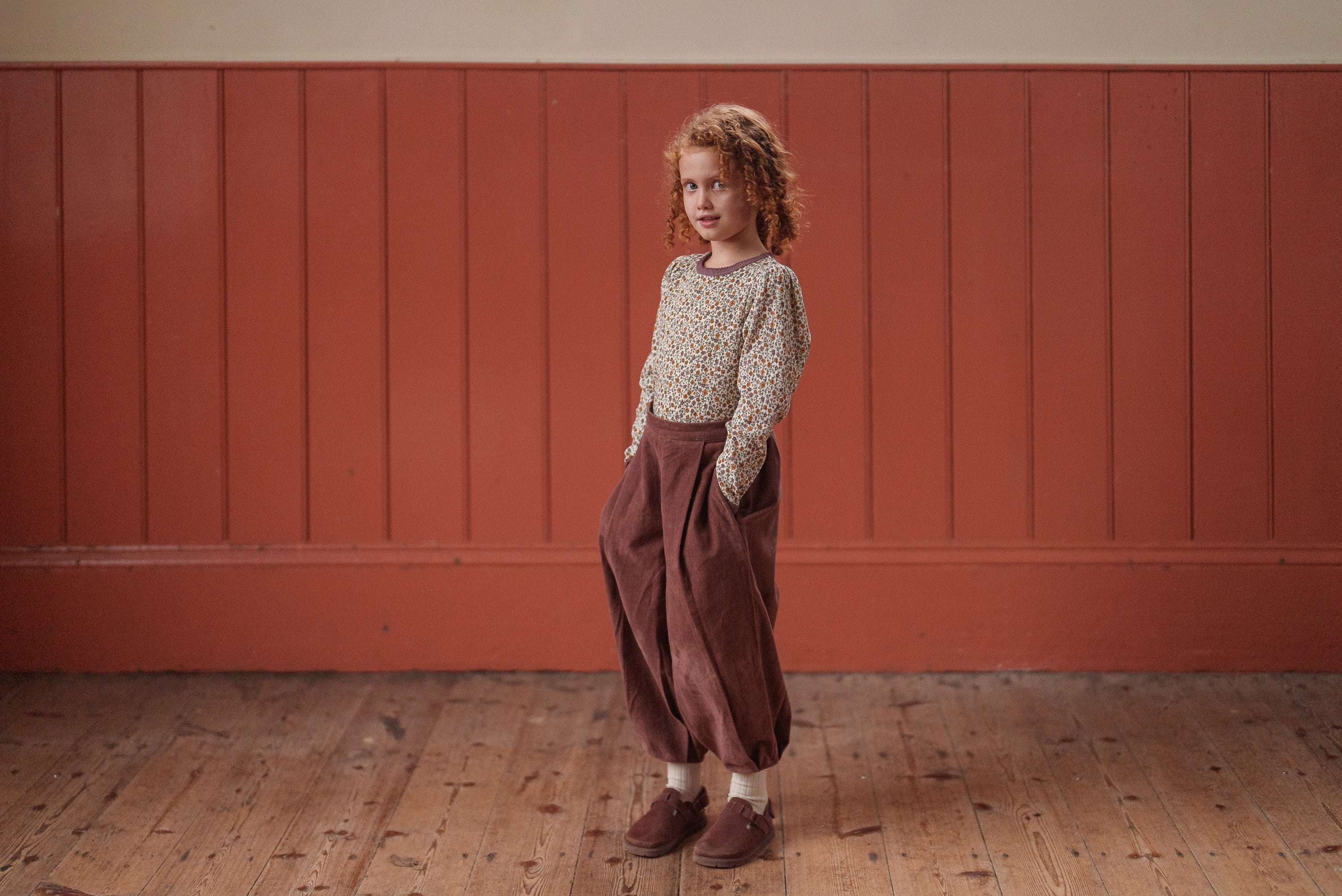 A young child stands indoors wearing natural, vintage-style children's clothing with loose, nutmeg-colored velvet trousers paired with a floral organic cotton top, embodying slow, ethical fashion.