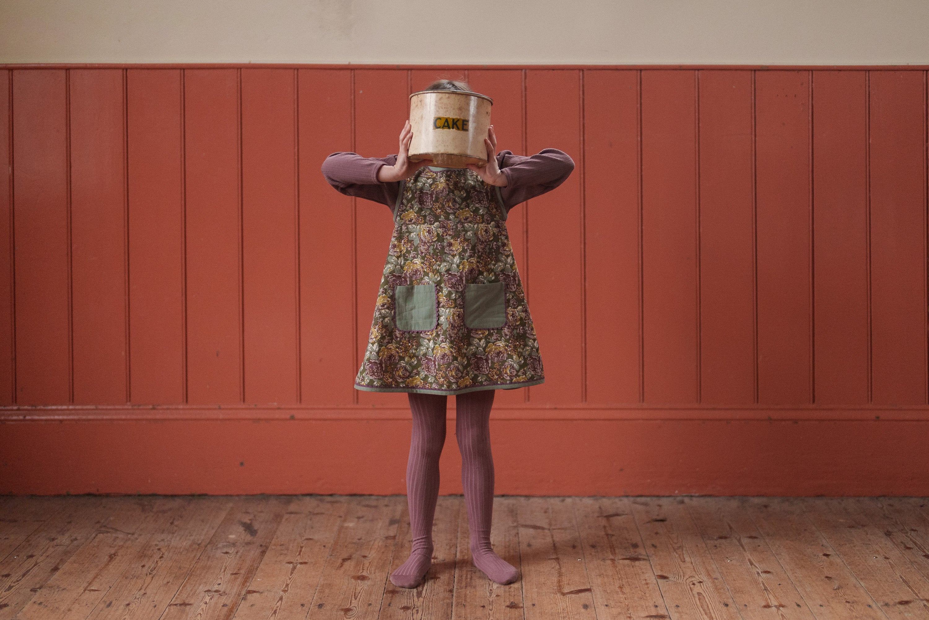 Child wearing a green floral organic cotton dress with green pockets, paired with vintage-style mauve tights, standing on a wooden floor against a muted red paneled wall holding a tin can over their face.