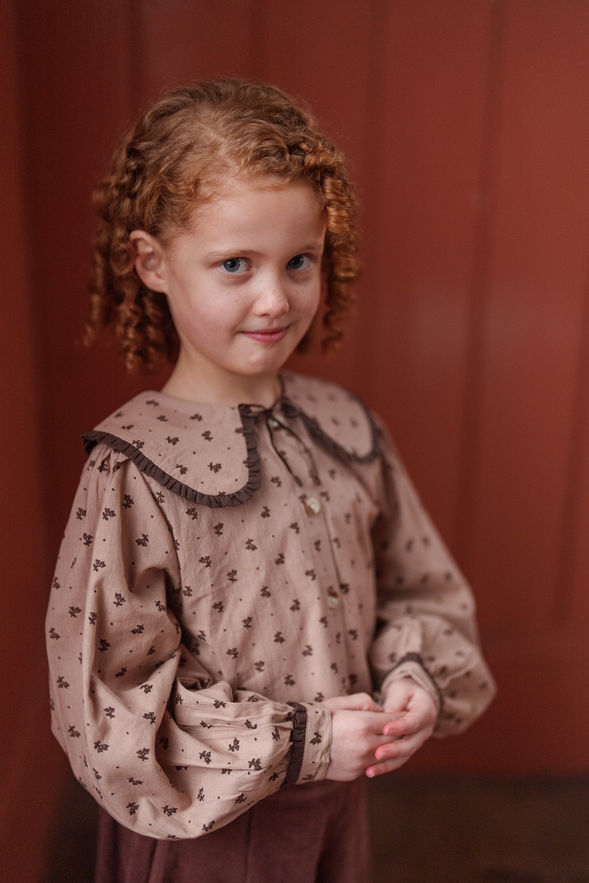Young girl wearing a beige organic cotton blouse with a rosehip floral pattern, featuring a vintage-inspired large collar and long puff sleeves, standing indoors.