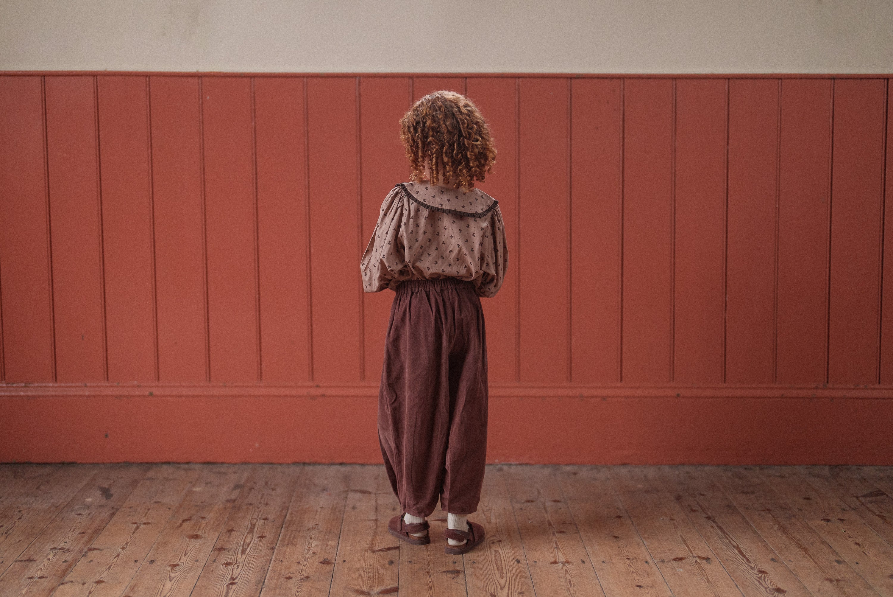 Child wearing vintage style, organic cotton clothing in warm, natural tones standing on wooden floor against a muted terracotta wall, showcasing sustainable kids wear.