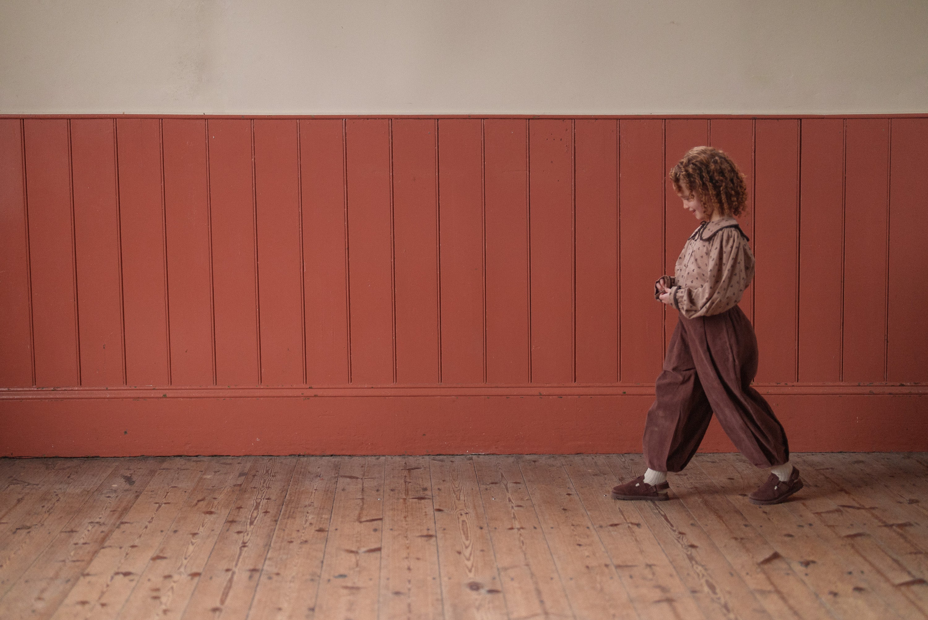 Young child with curly hair wearing vintage style kids clothing in earthy tones, including a floral organic cotton blouse and loose trousers, walking indoors on wooden floor.