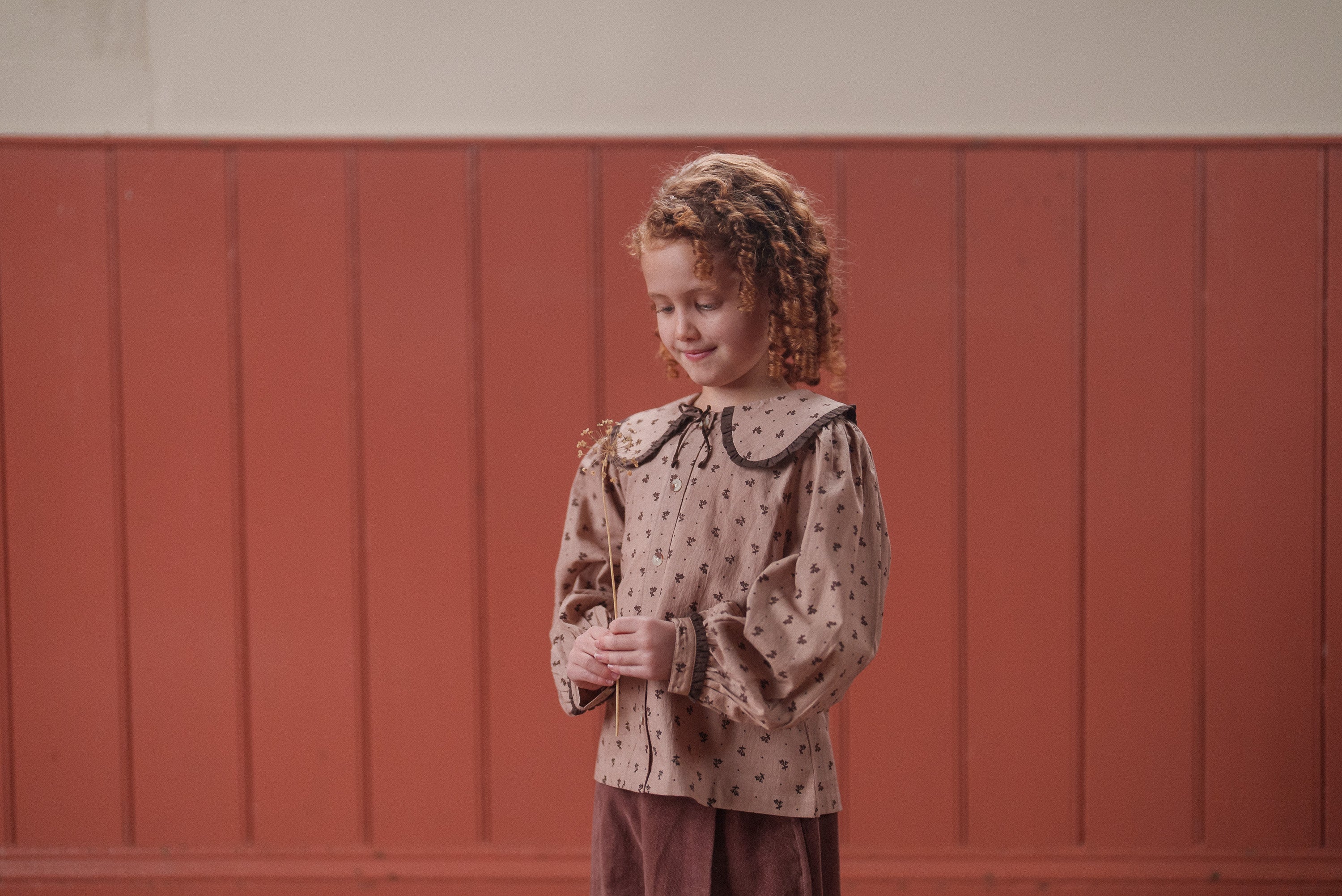 Young girl wearing a beige organic cotton blouse with a rosehip floral pattern and vintage-inspired collar, standing against a warm wooden paneled background in sustainable kids clothing.
