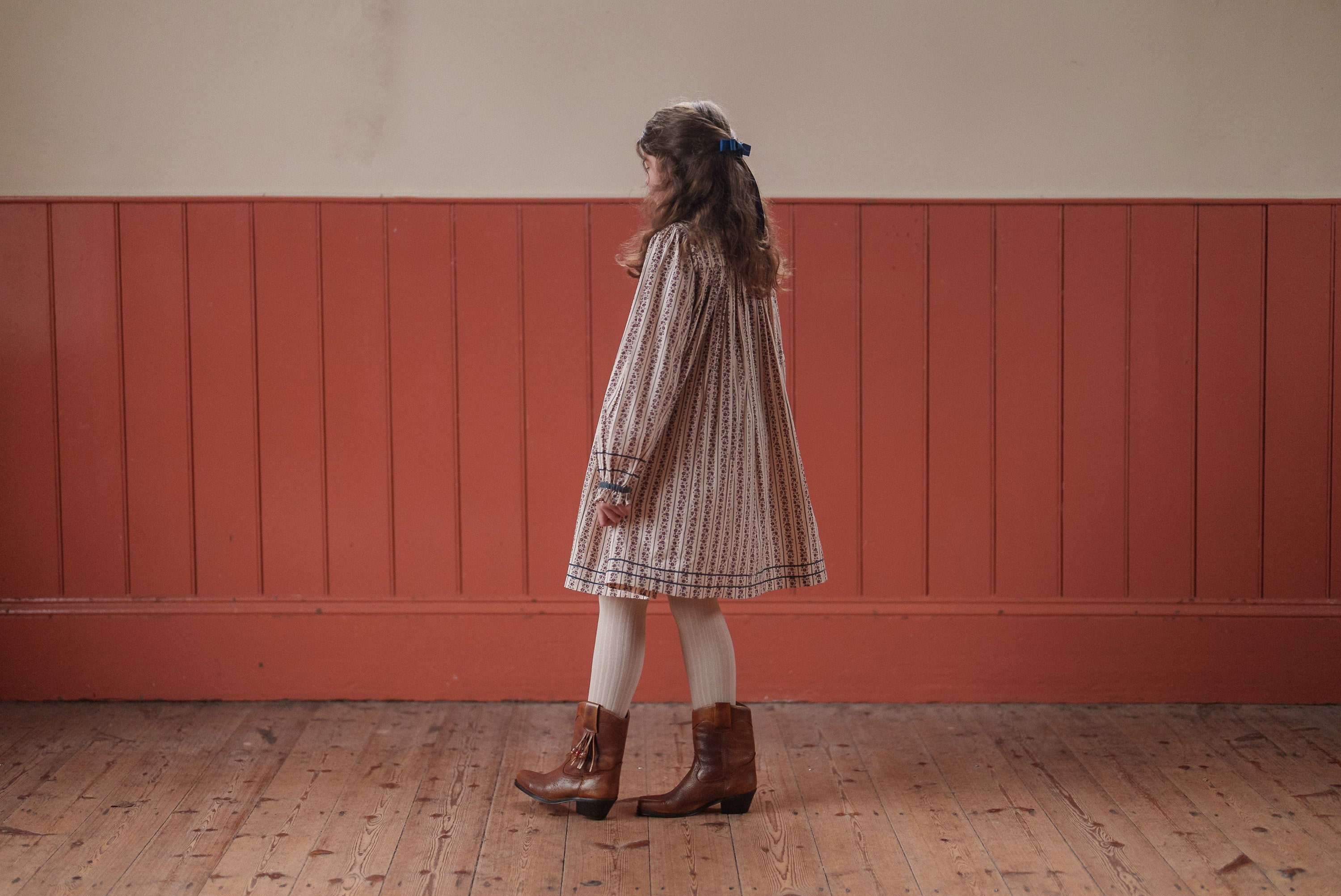Young girl wearing an organic cotton smocked dress with a vintage style, paired with tights and brown boots, standing on a wooden floor in a warm, natural setting.