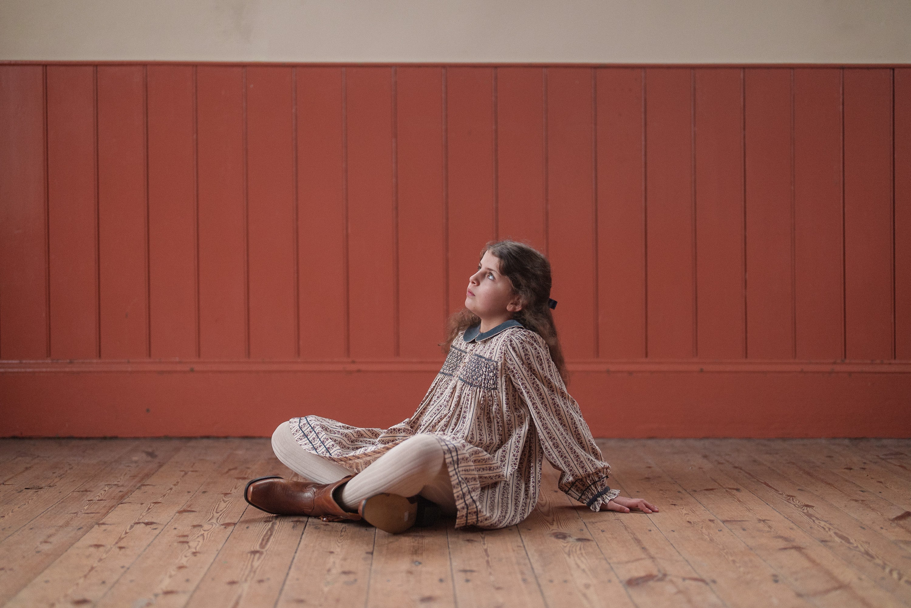 Young girl wearing a vintage style organic cotton dress sitting on a wooden floor in a natural, softly lit room, reflecting a slow and ethical children’s clothing brand.