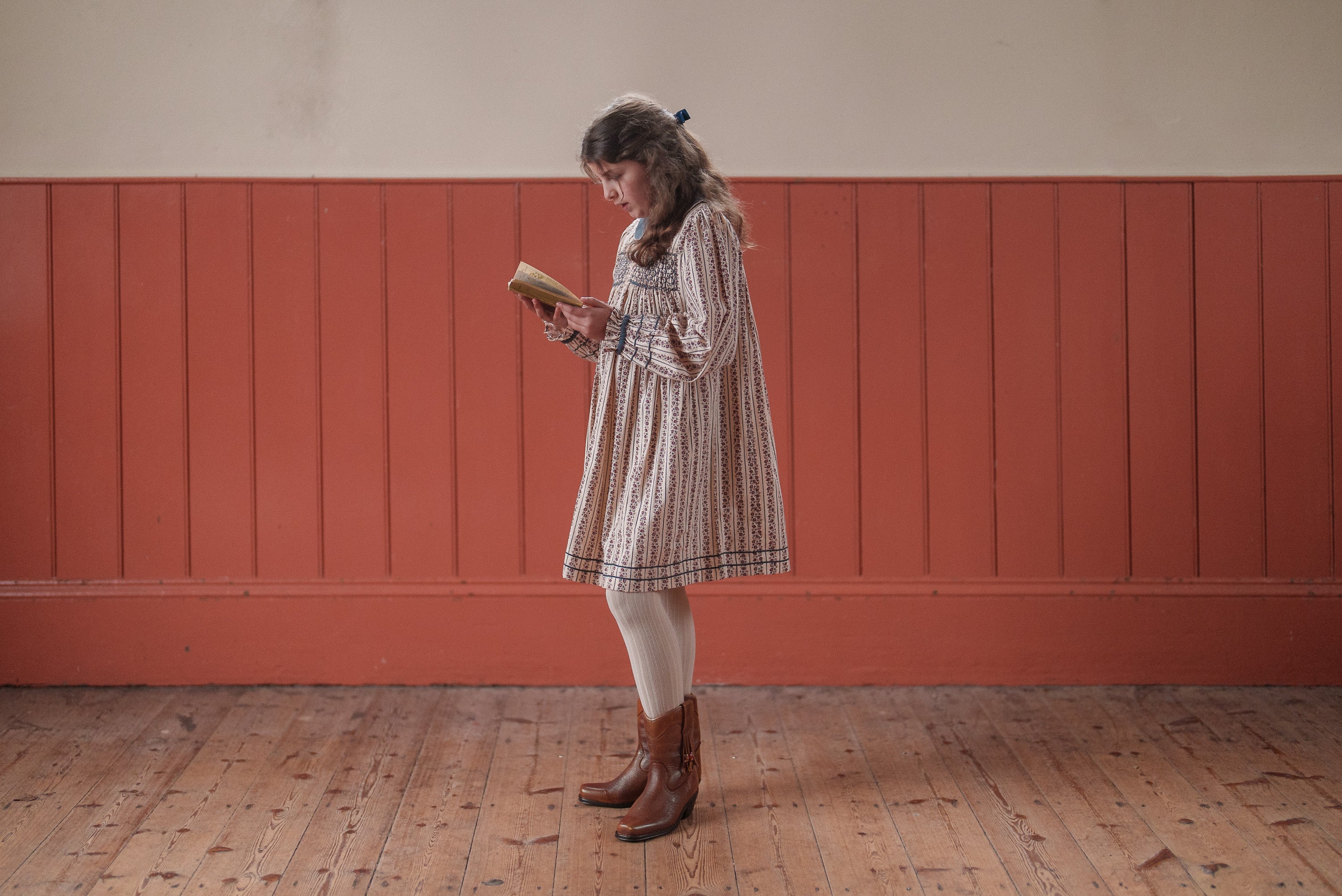 Young girl wearing an organic cotton vintage-style dress with Mallory floral smocking, paired with tights and brown boots, reading a book in a warm, natural setting.